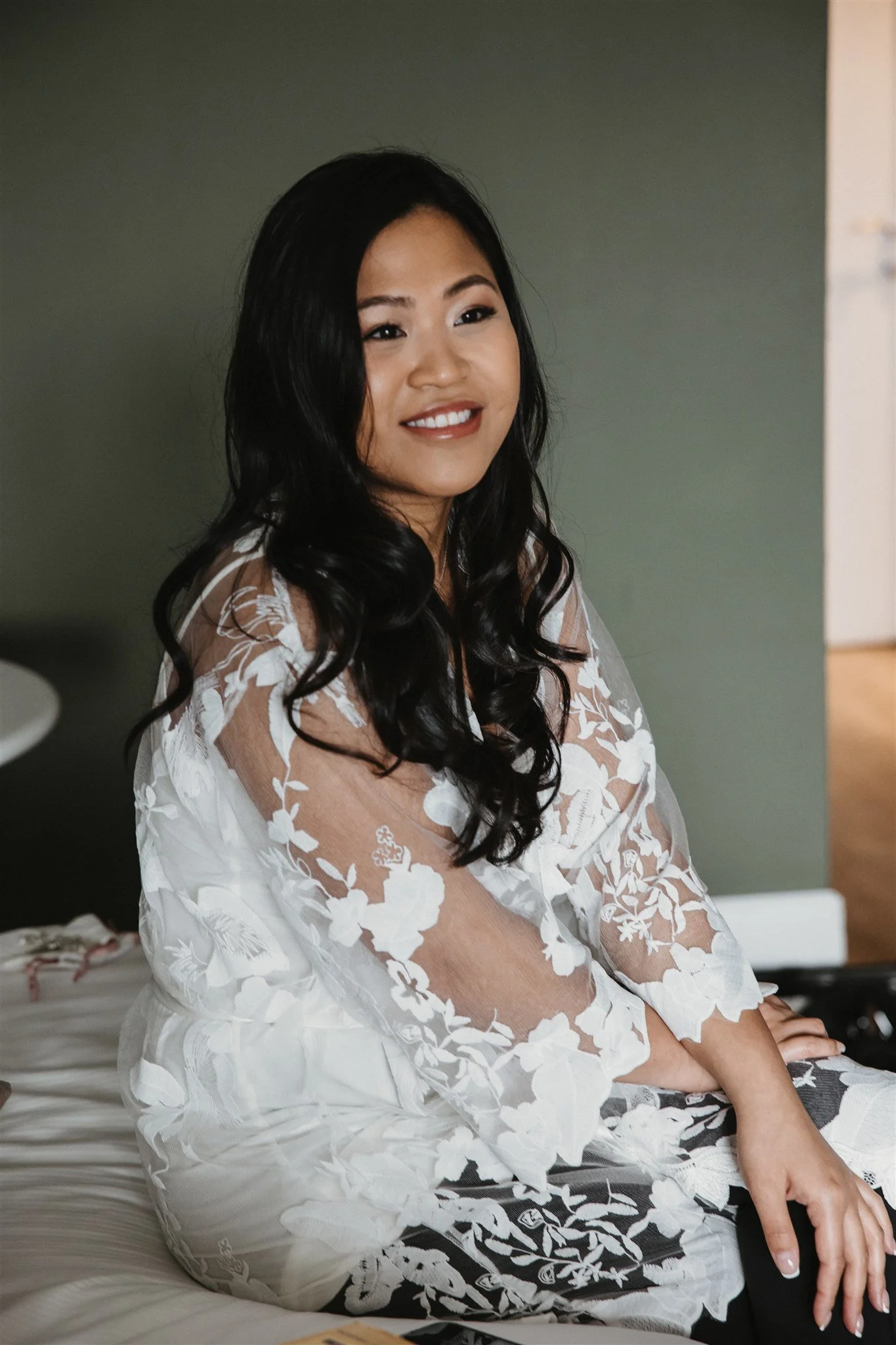 A woman with black hair styled in loose curls, smiling and sitting on a bed, wearing a white lace dress with floral embroidery.