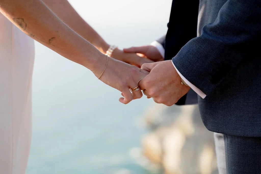 Close-up of a couple holding hands during a wedding, with the ocean and rocky cliffs blurred in the background.