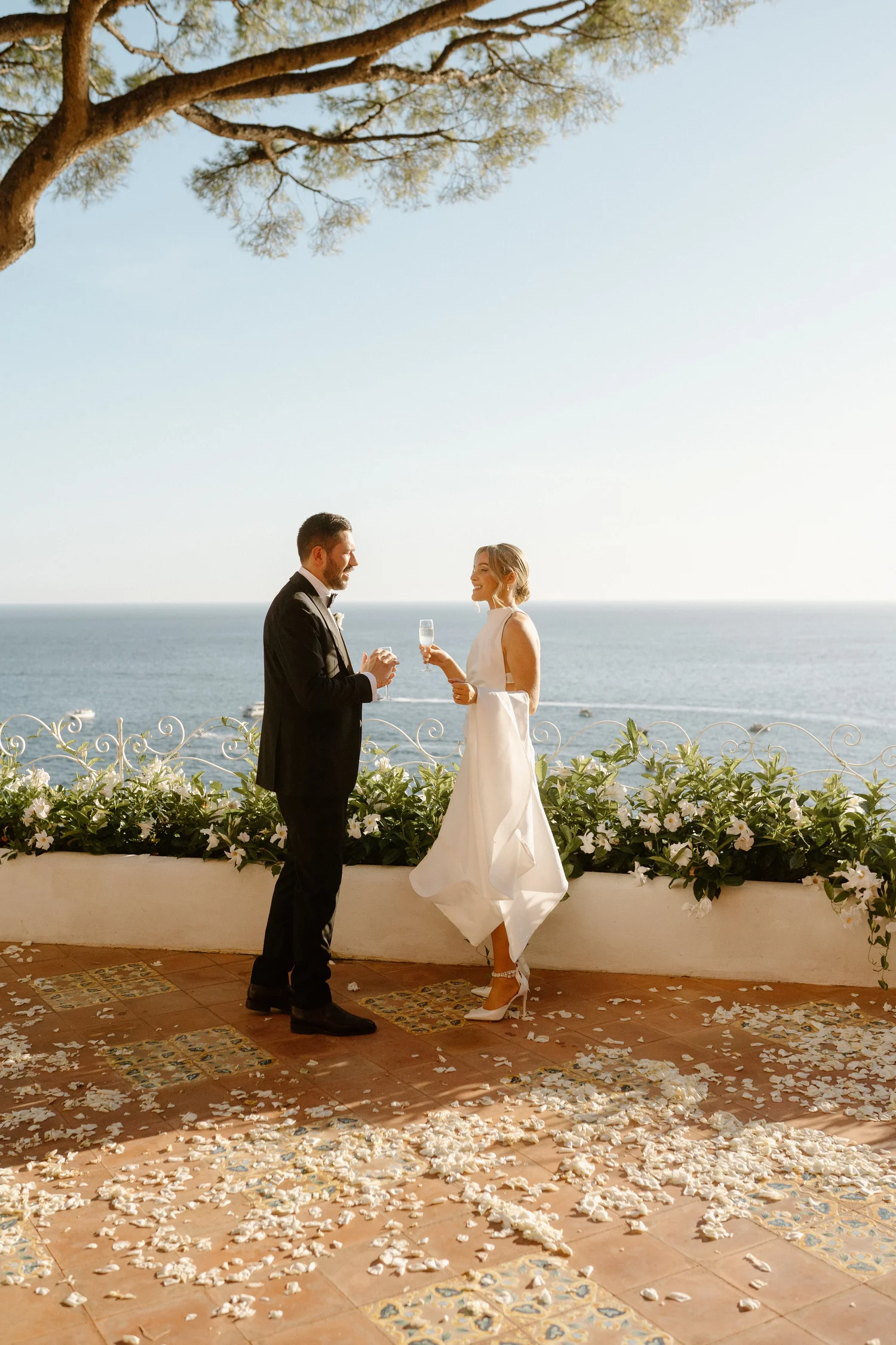 A wedding ceremony taking place outdoors by the ocean, with a bride and groom standing on a tiled floor scattered with flower petals, under a large tree, holding glasses and smiling at each other.