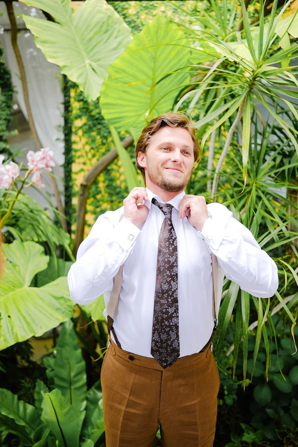 A man with light brown hair and a beard adjusting his tie in a lush green indoor garden. He is wearing a white dress shirt, a patterned tie, and brown pants with suspenders.