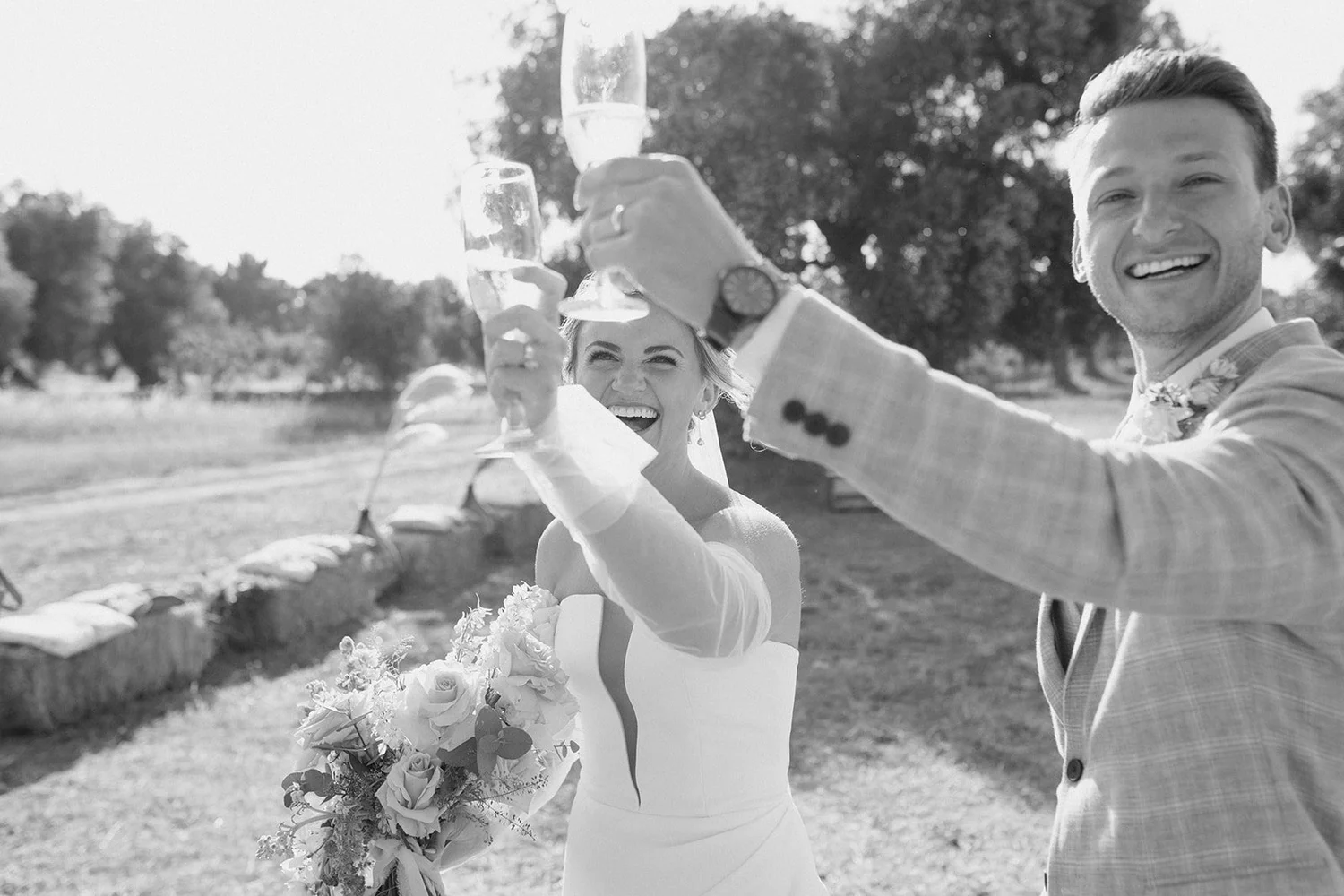 A black-and-white photo of a smiling bride and groom holding up glasses for a toast outdoors, with trees in the background.