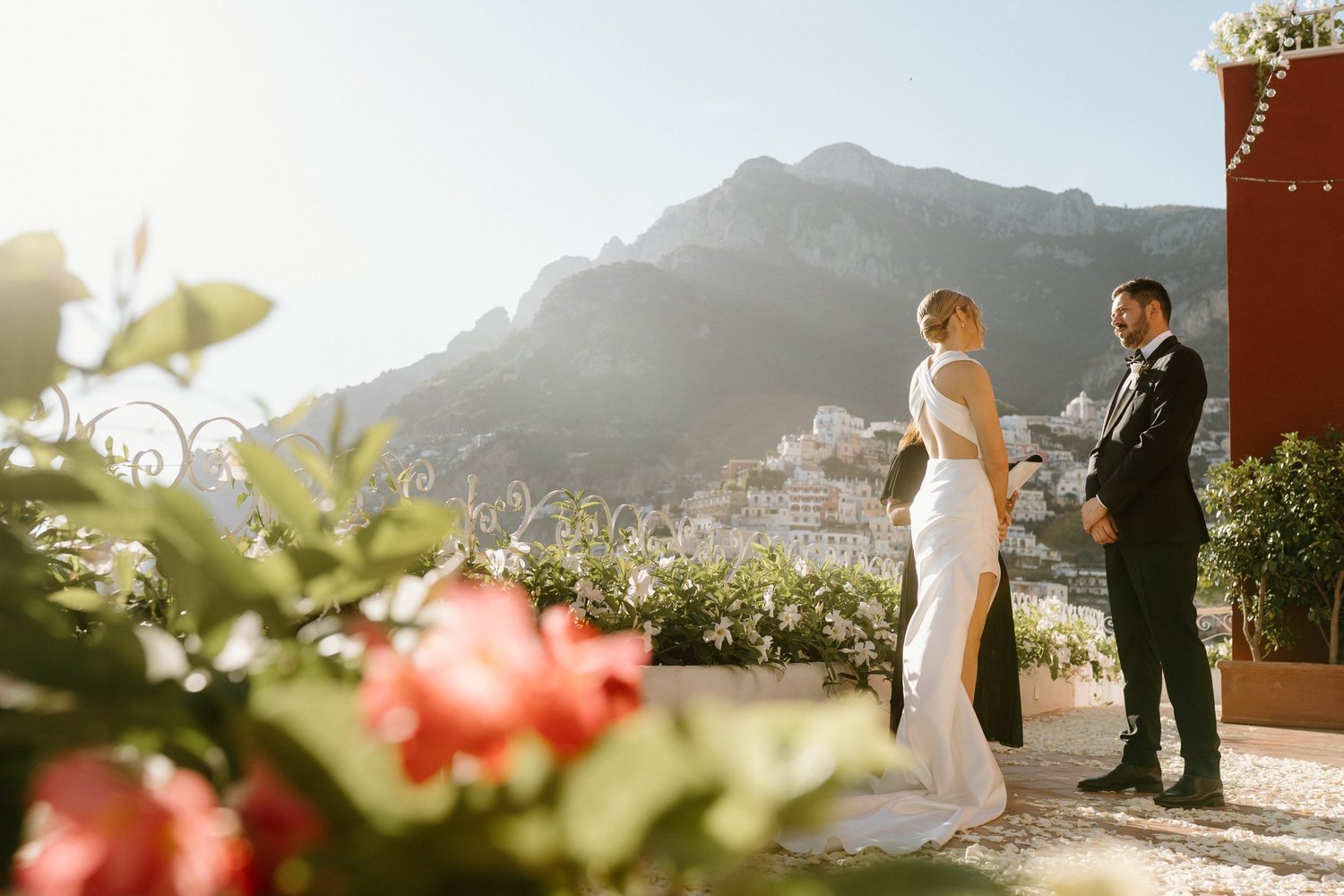 A bride and groom standing outdoors during their wedding ceremony with mountains in the background, flowers in the foreground, and the sunset casting a warm light.