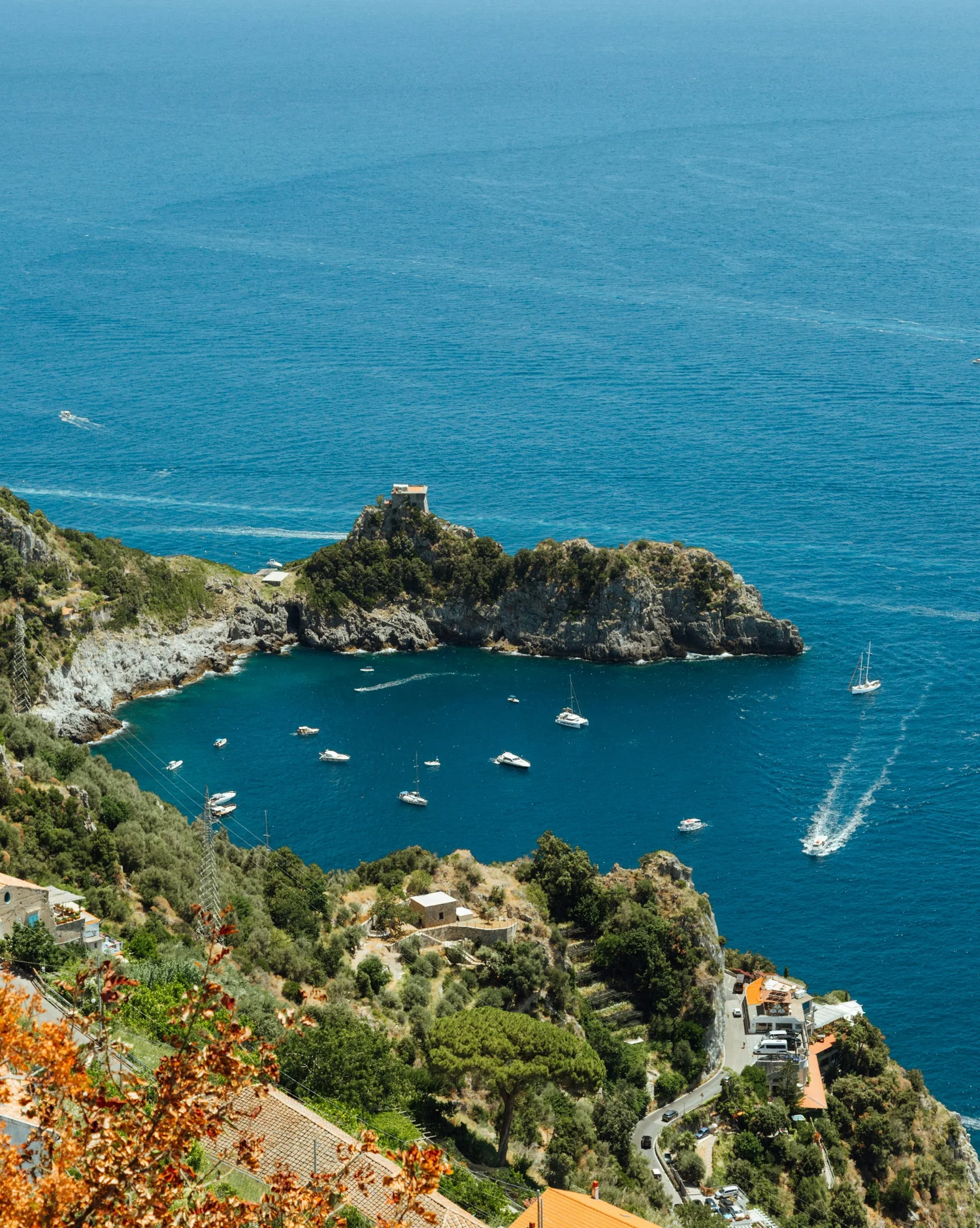 Coastal view with turquoise water, boats, and colorful houses on a hillside with trees and greenery.