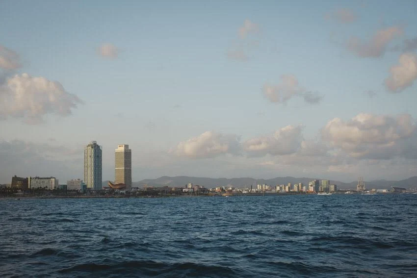 City skyline with tall buildings along the waterfront under a partly cloudy sky, with a body of water in the foreground.