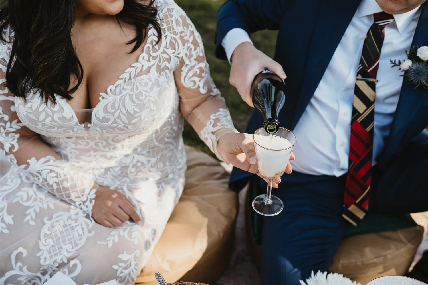 A woman in a white, lace wedding dress and a man in a suit pour champagne into a flute at an outdoor celebration.