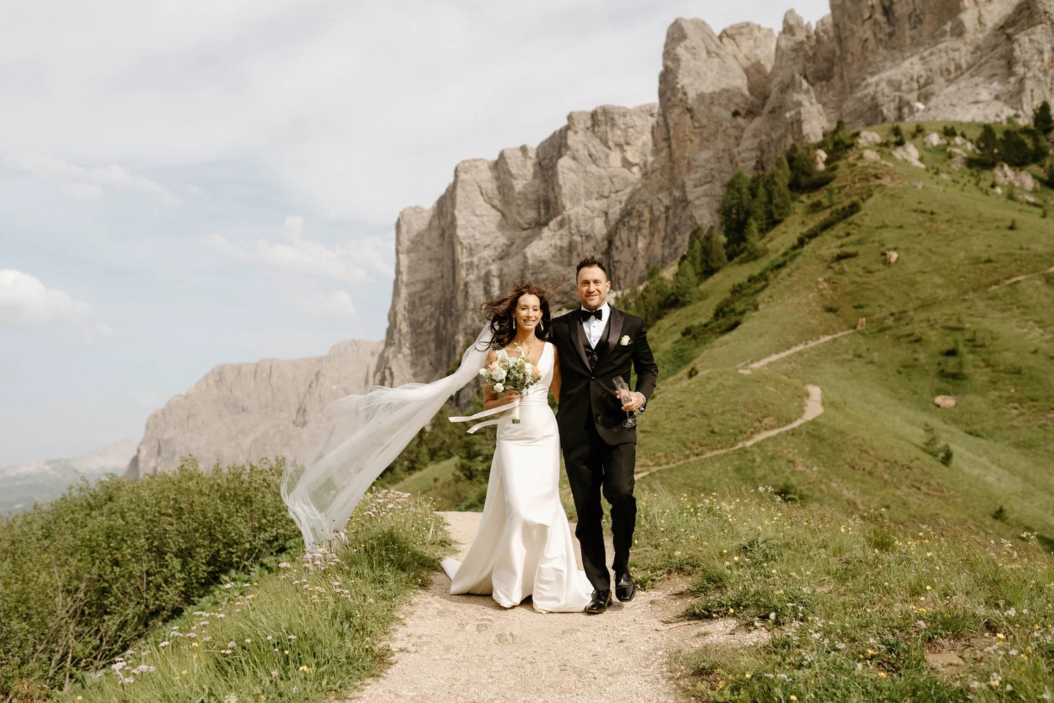 A newlywed couple walking down a trail on a grassy hillside with rocks and mountains in the background. The bride is wearing a white wedding dress with a long veil and holding a bouquet, while the groom is in a black tuxedo. They are smiling and hold