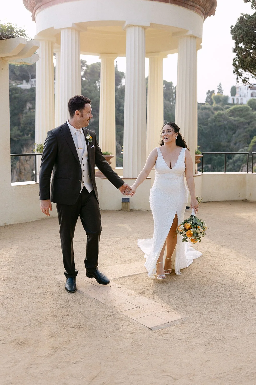 A bride and groom holding hands and smiling at each other outdoors during their wedding, with classical columns and lush greenery in the background.