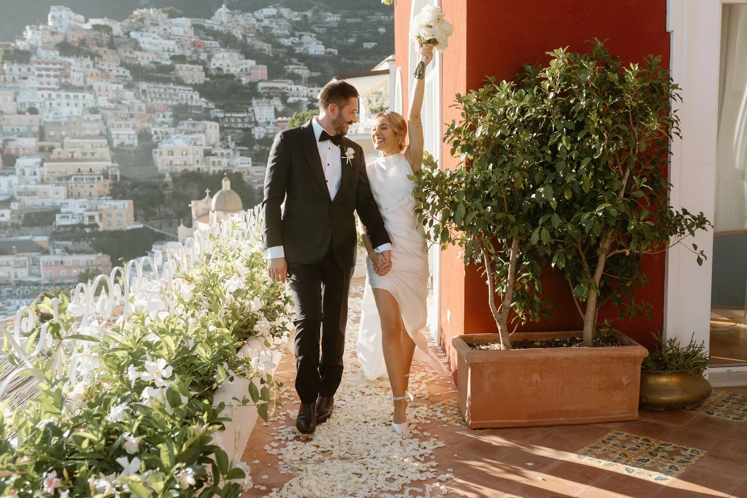 A happy bride and groom holding hands, walking on a flower petal-covered pathway on a balcony overlooking a hillside with colorful buildings. The bride is smiling with her arm raised, holding a bouquet, and the groom is in a tuxedo with a bow tie. Bright sunlight illuminates the scene.