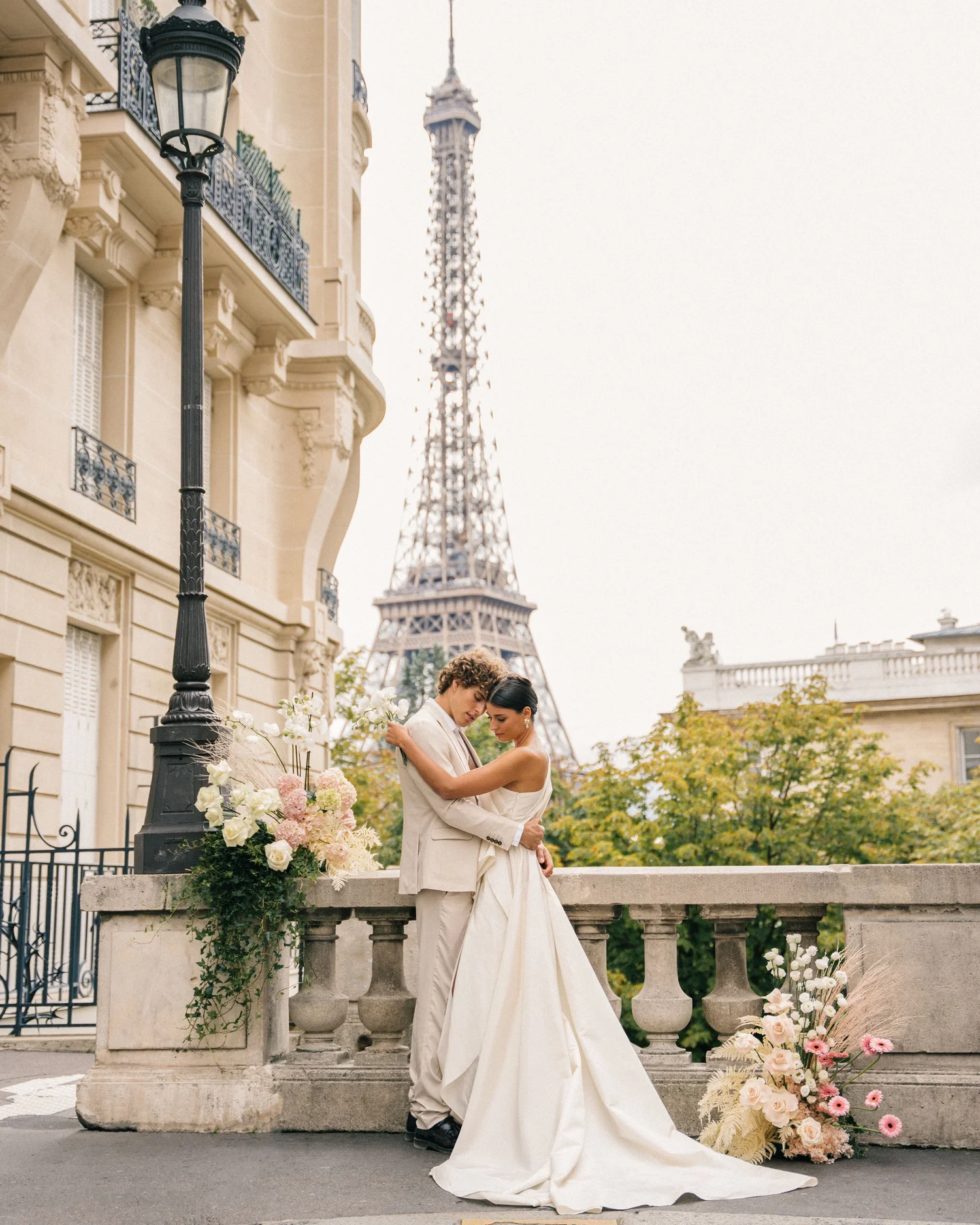 A couple dressed in wedding attire embraces on a stone balcony decorated with white and pink flowers, with the Eiffel Tower in the background in Paris, France.
