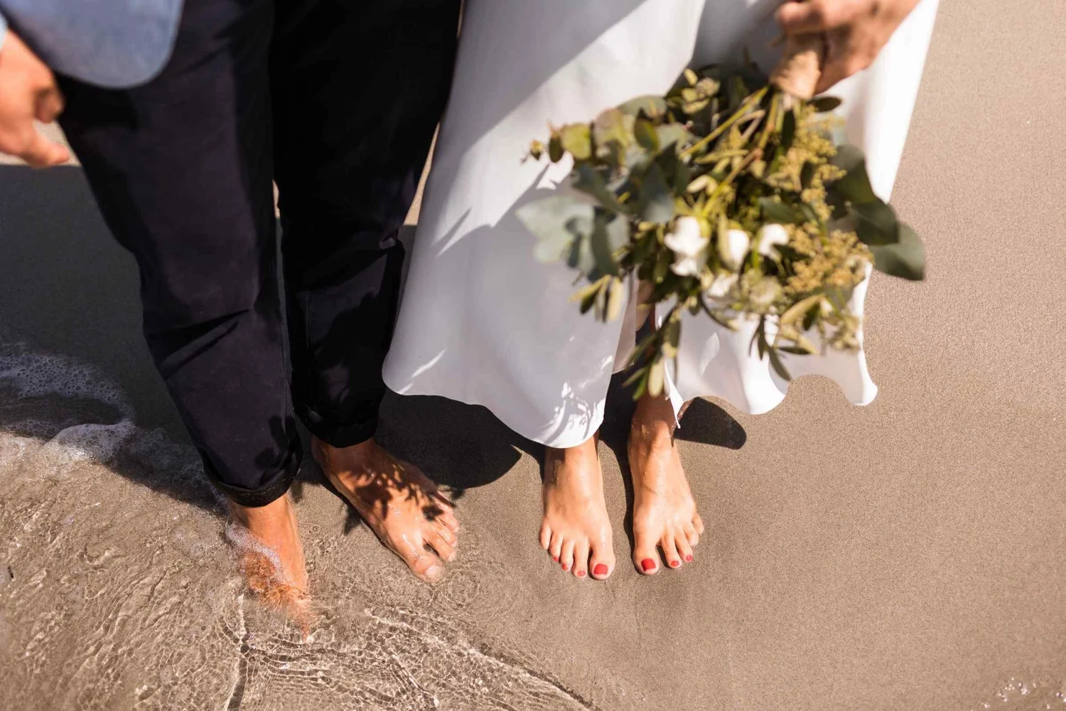 Two people stand barefoot in the shallow water at the beach, with a white tablecloth on a small table and a bouquet of flowers placed on it.