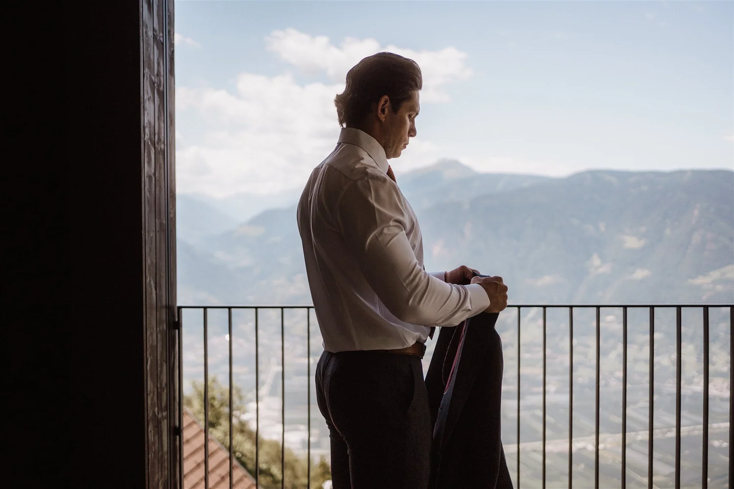 A man in a white shirt and black pants standing on a balcony with mountain views, looking down at his phone.