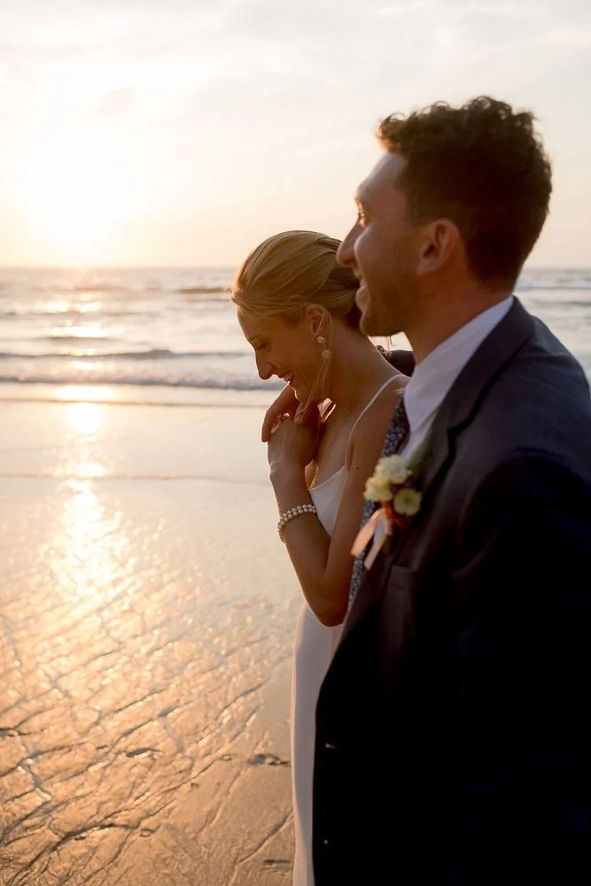 A couple in wedding attire standing on the beach at sunset, sharing a joyful moment.