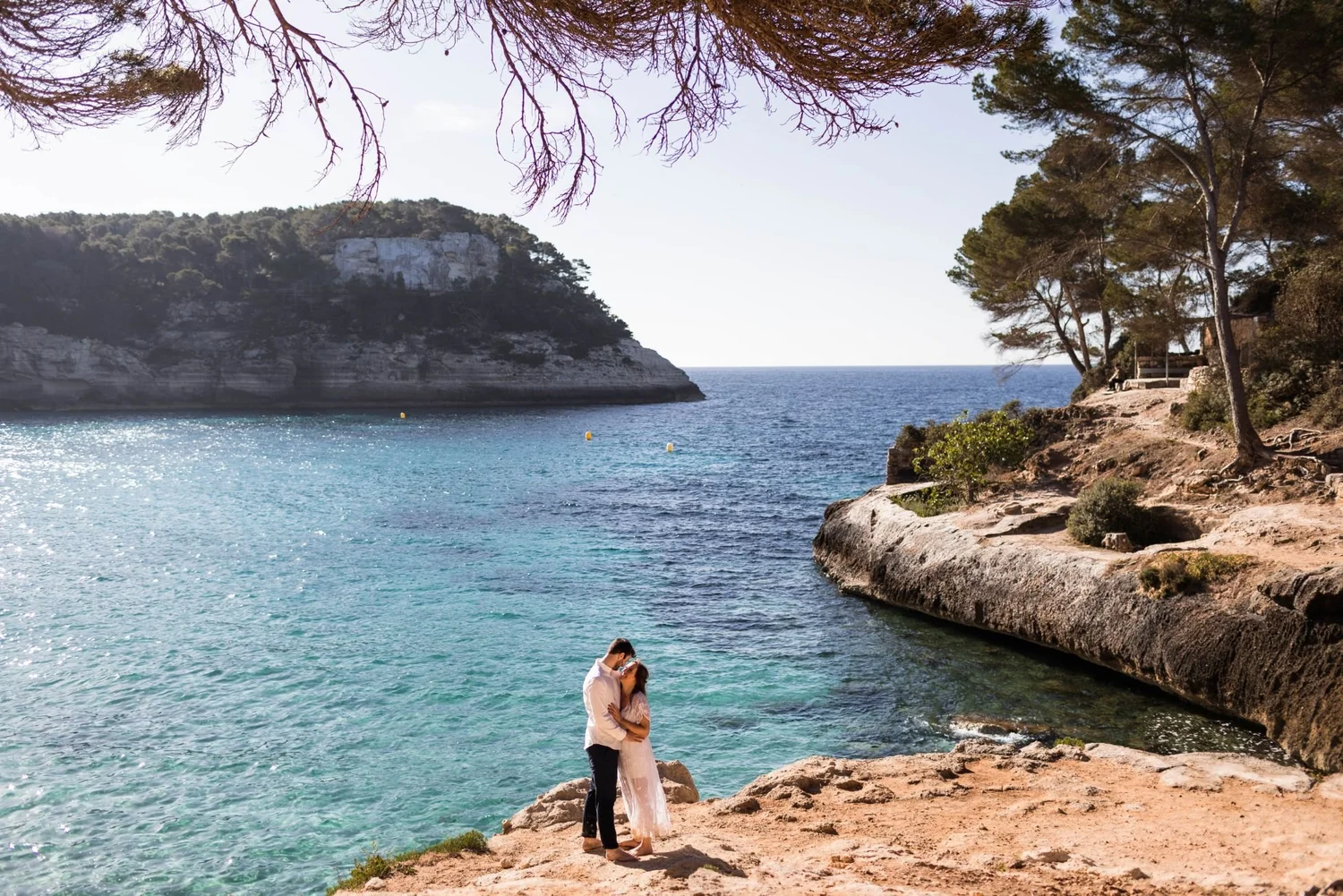 A couple embraces on a rocky beach by the sea, with cliffs and trees in the background. The man wears dark pants and a white shirt, while the woman wears a light-colored dress.