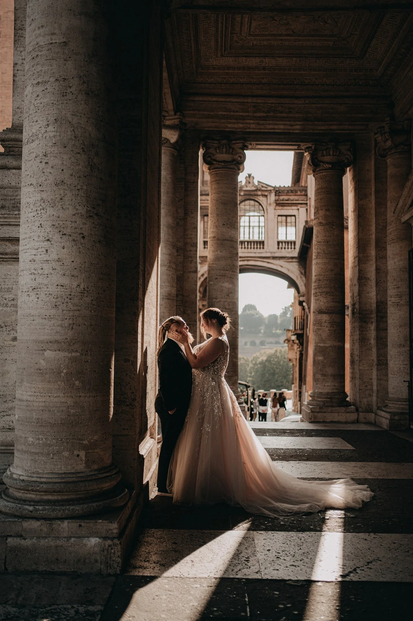 A bride and groom standing close together under an archway with large stone columns, with city buildings and people visible in the background, illuminated by warm sunlight.