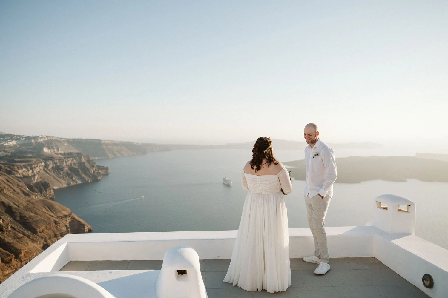 A couple stands in front of a scenic ocean view with cliffs, during their wedding ceremony. The bride wears a white off-shoulder dress with long hair, and the groom wears a white shirt and light trousers. The setting appears to be in a sunny, coastal