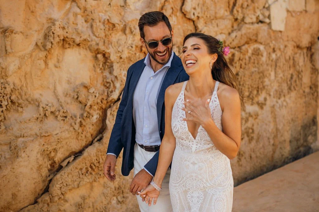 A happy couple with light skin walking outdoors near a rocky wall, dressed in wedding attire, smiling and holding hands.