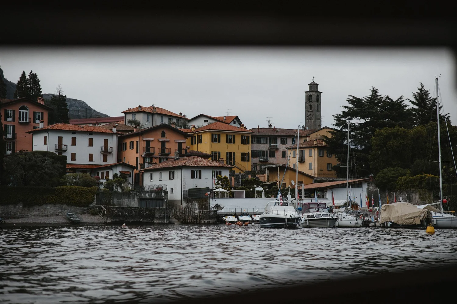 A waterfront scene with a small marina and colorful buildings, trees, and hills in the background under a cloudy sky.