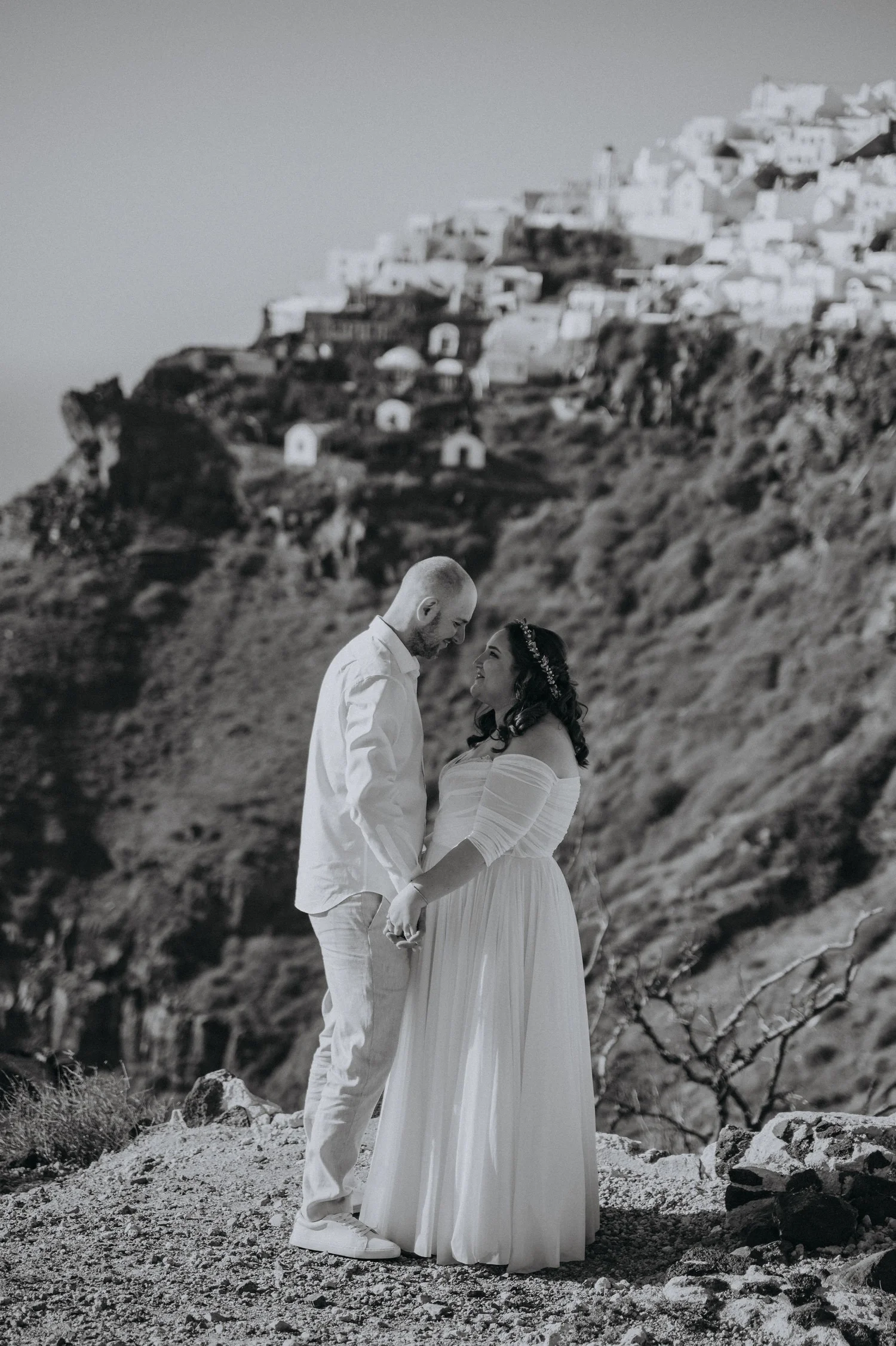 A couple stands on rocky terrain with a hillside village in the background. The woman is wearing a white off-the-shoulder dress and headpiece, and the man is in a white shirt and pants. They are holding hands and looking at each other, appearing to b