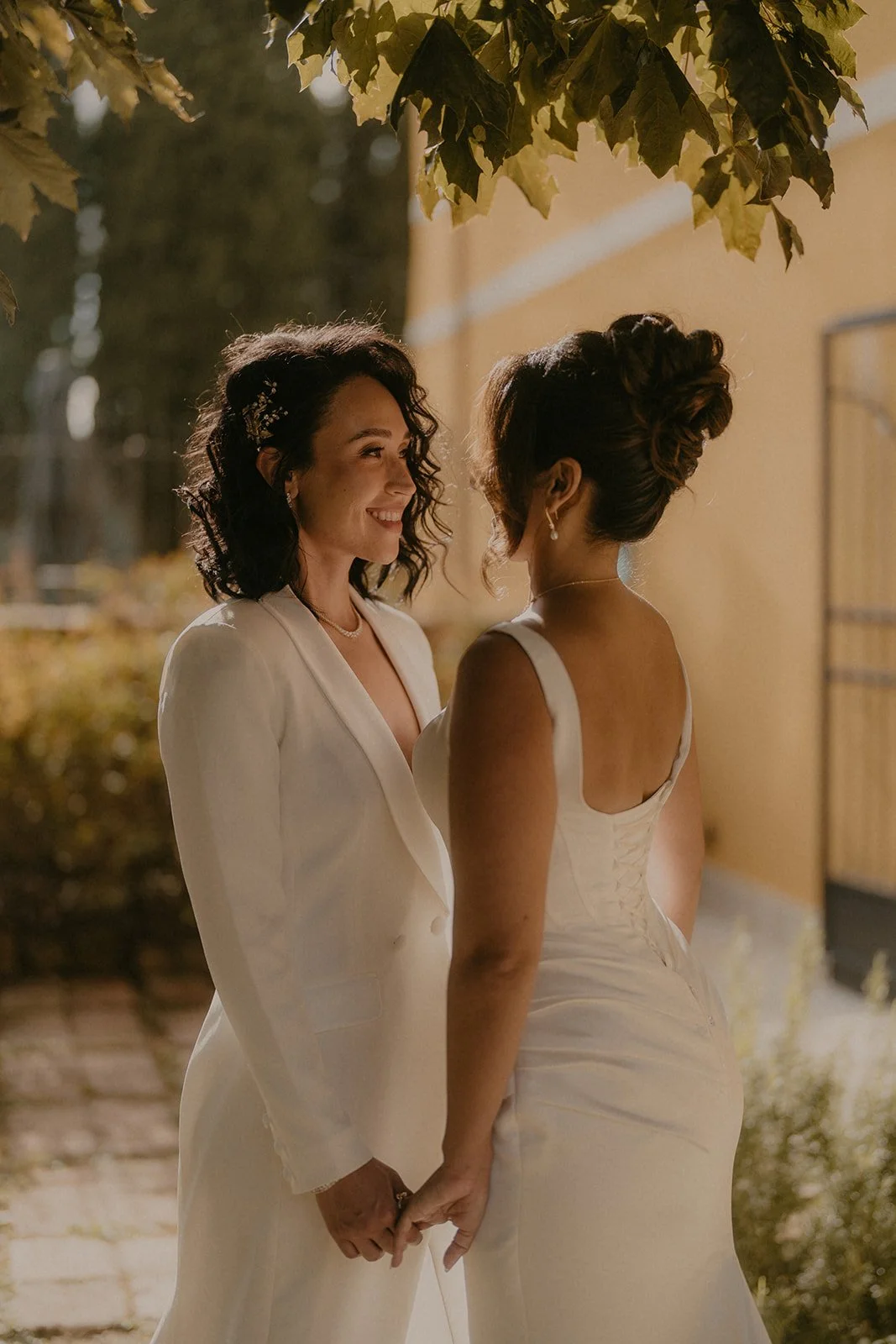Two women in wedding dresses holding hands and smiling at each other outdoors at sunset.