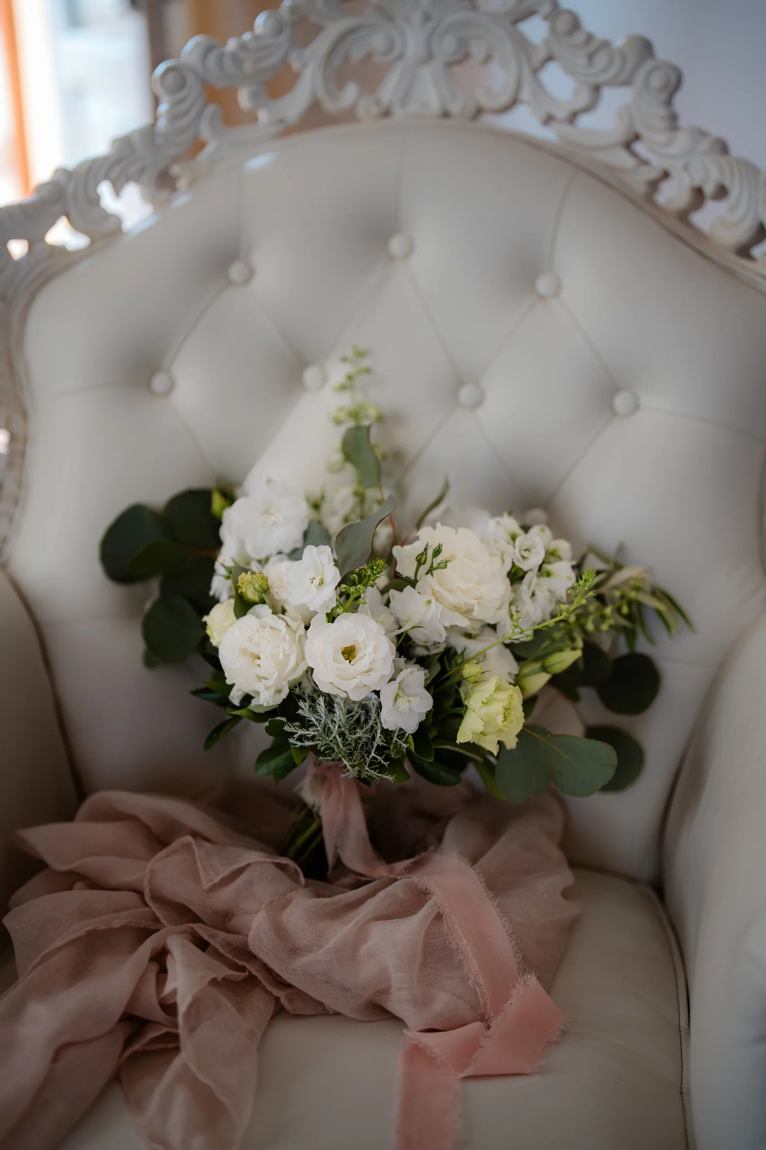 A bouquet of white flowers with green leaves, tied with a pink ribbon, resting on a cushioned armchair with ornate white detailing.