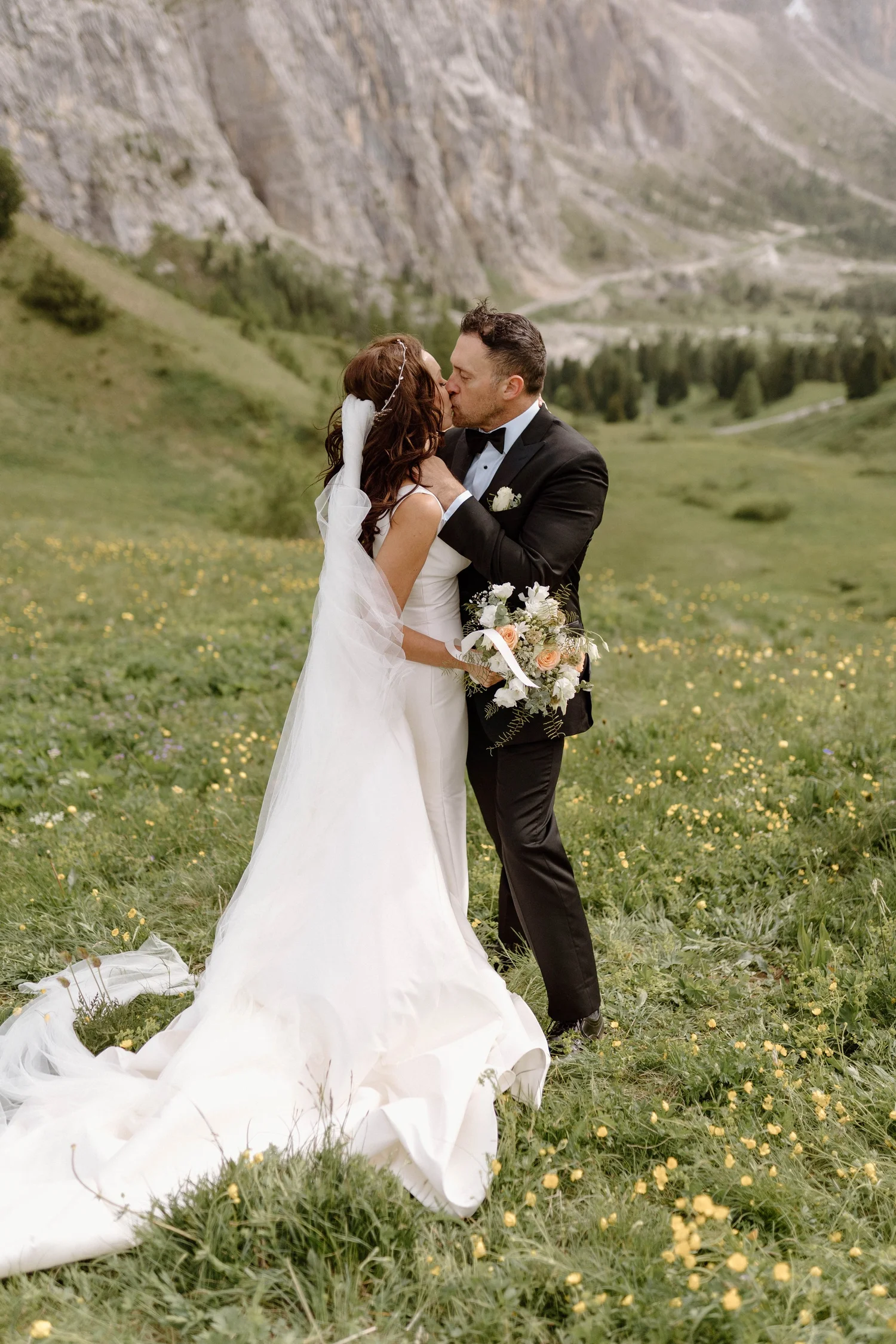 A bride and groom sharing a kiss outdoors in a green field with yellow flowers, surrounded by mountains and trees.