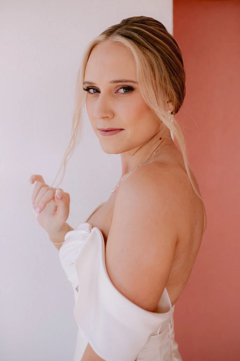 A woman with blonde hair styled in an updo with loose strands, smiling slightly, wearing a white off-shoulder dress against a pink and white background.