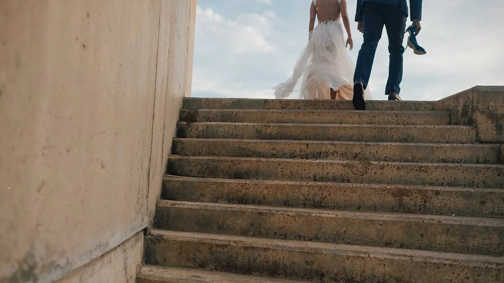 Bride and groom walking up outdoor concrete stairs, back view, wedding dress and suit, cloudy sky