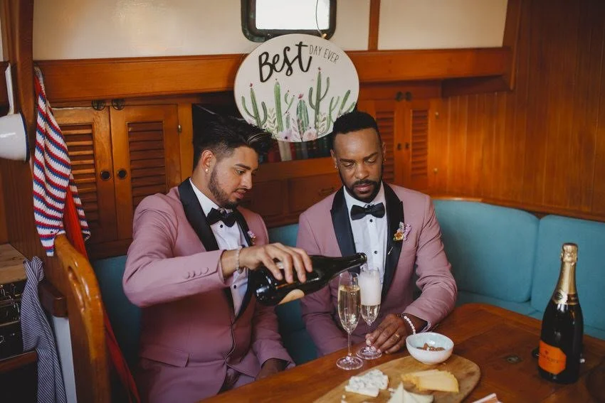 Two men in pink tuxedos with black lapels sitting at a wooden table with blue cushioned seats, pouring champagne into glasses, with a basket of snacks and a bottle of champagne on the table, in a wood-paneled room decorated with a cactus-themed sign
