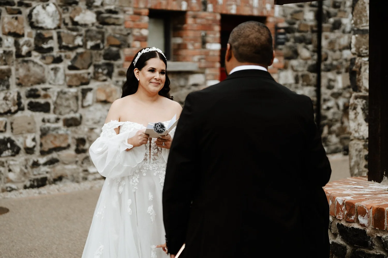 A bride standing in front of a man during a wedding ceremony outdoors with a stone and brick wall in the background.