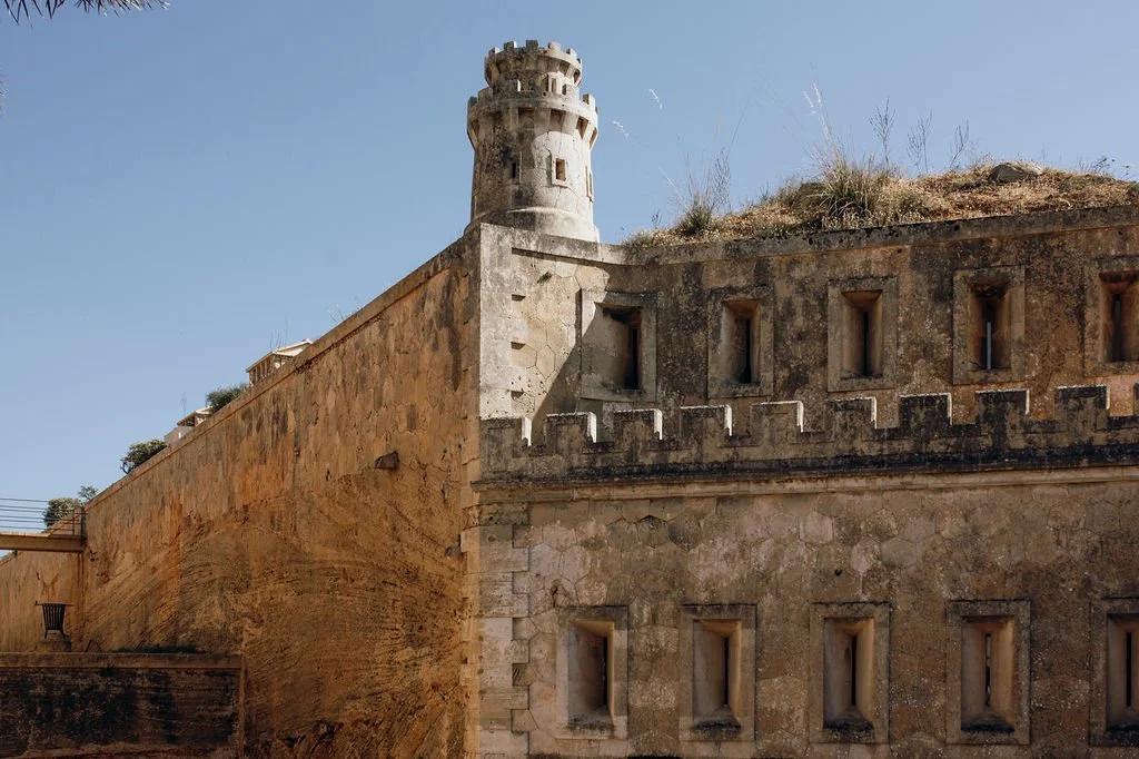 An old stone fortress with a round tower and narrow windows, built with beige and brown stones, under a clear blue sky.