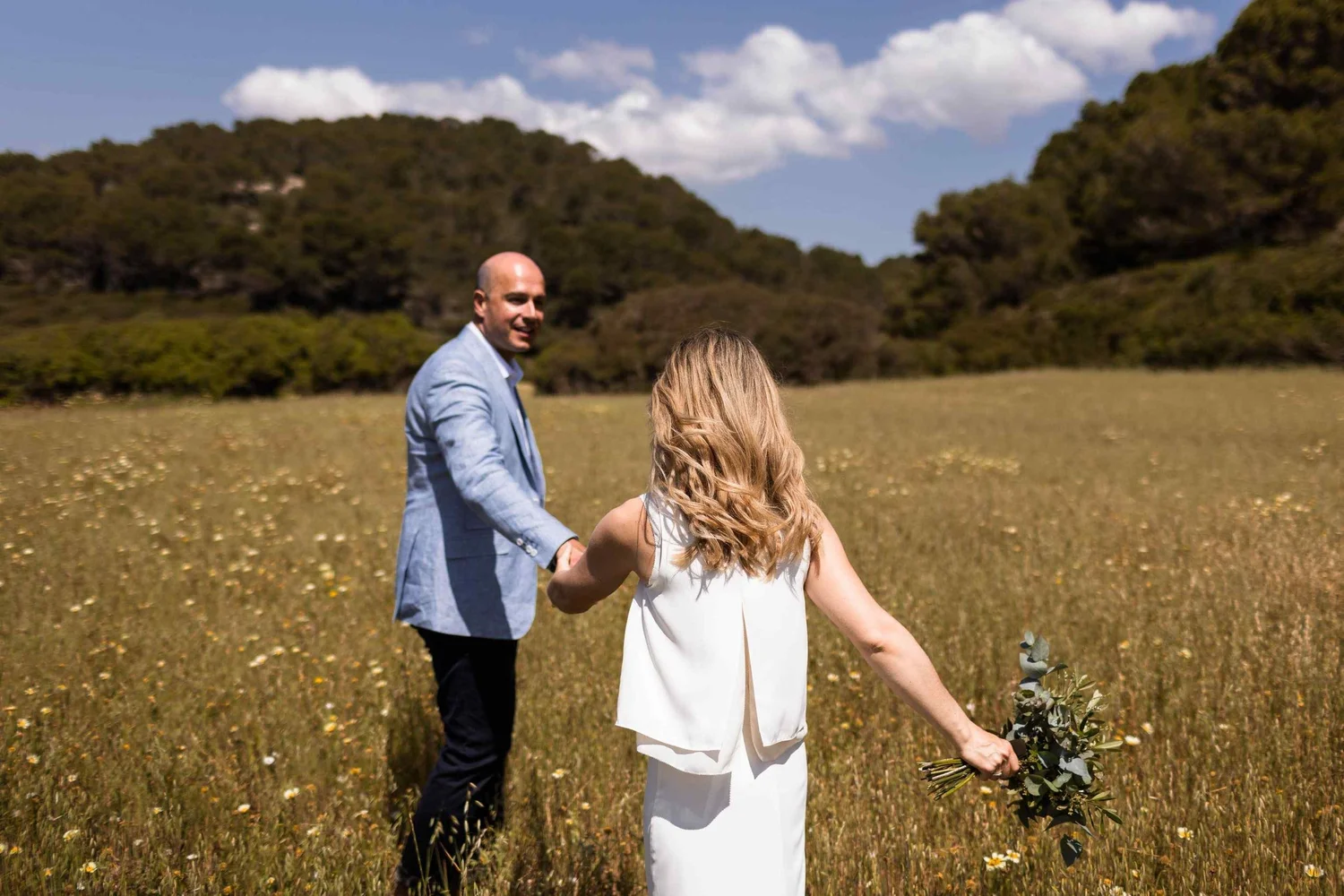 A man and woman holding hands in a field of yellow flowers with hills and a blue sky in the background.
