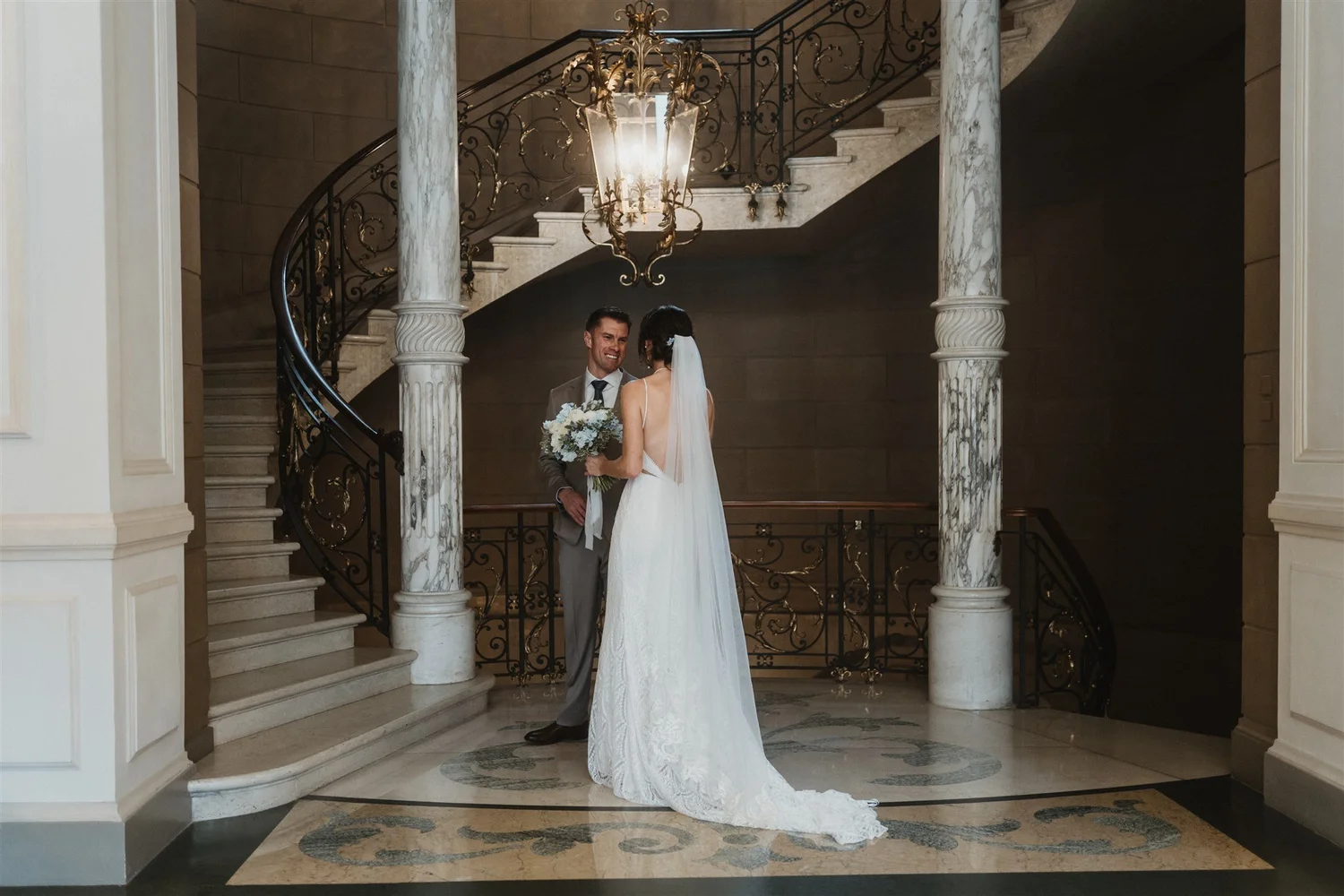 A bride and groom stand on a staircase landing inside a grand building, sharing a moment. The bride is in a white gown with a long train and veil, holding a bouquet. The groom is in a light gray suit. A large, ornate chandelier hangs above them, and