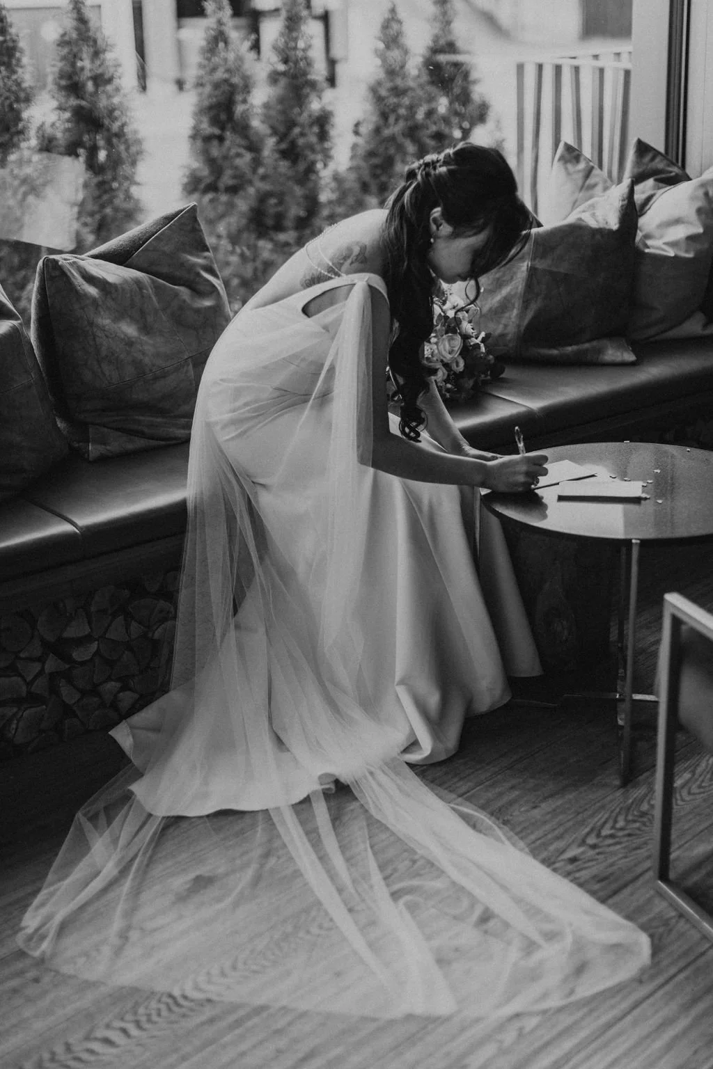 A bride in a wedding dress signing a document while leaning over a small table inside, with a sofa and cushions behind her and trees outside a window.