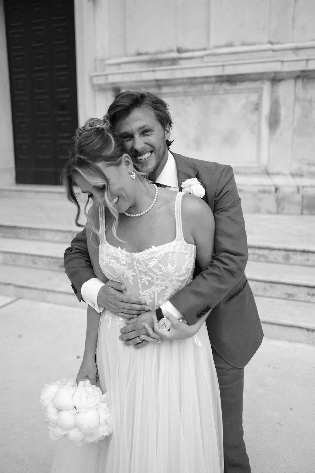 A smiling bride and groom embrace on wedding steps, with the groom in a suit and the bride in a wedding dress holding a bouquet.