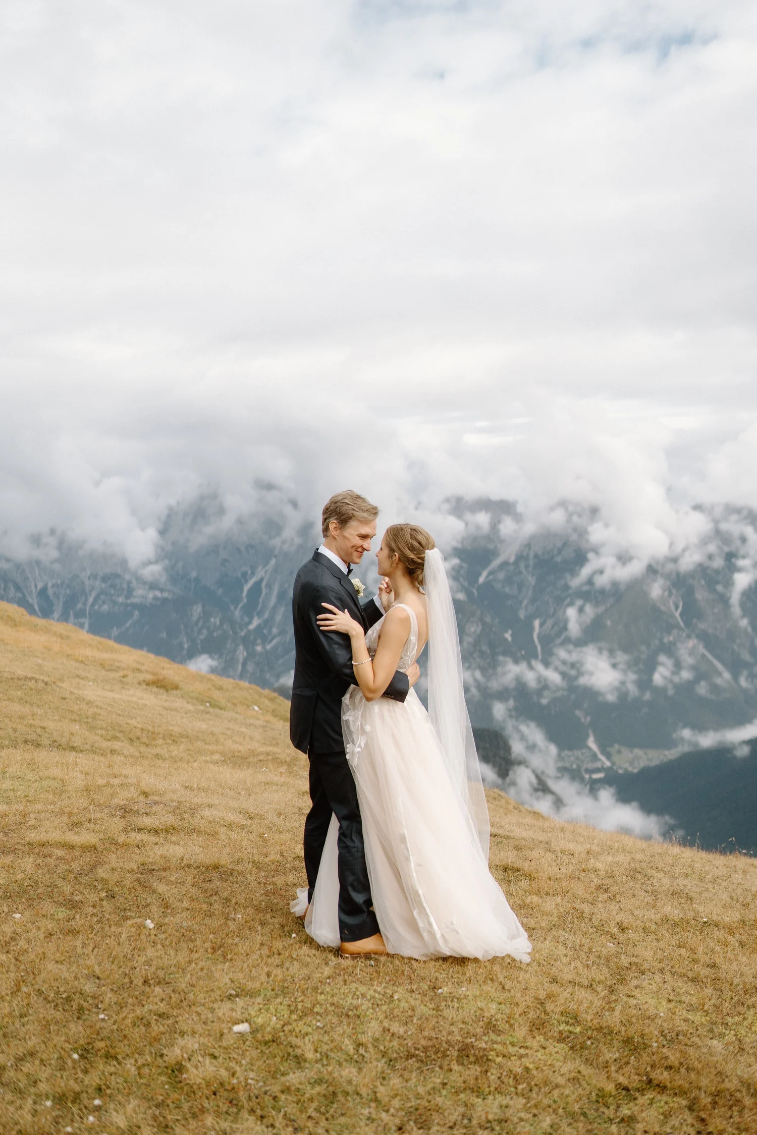 A bride and groom standing face to face on a grassy hill with mountains and cloudy sky in the background. The bride wears a white gown and veil, and the groom wears a dark suit. They are gazing at each other lovingly.