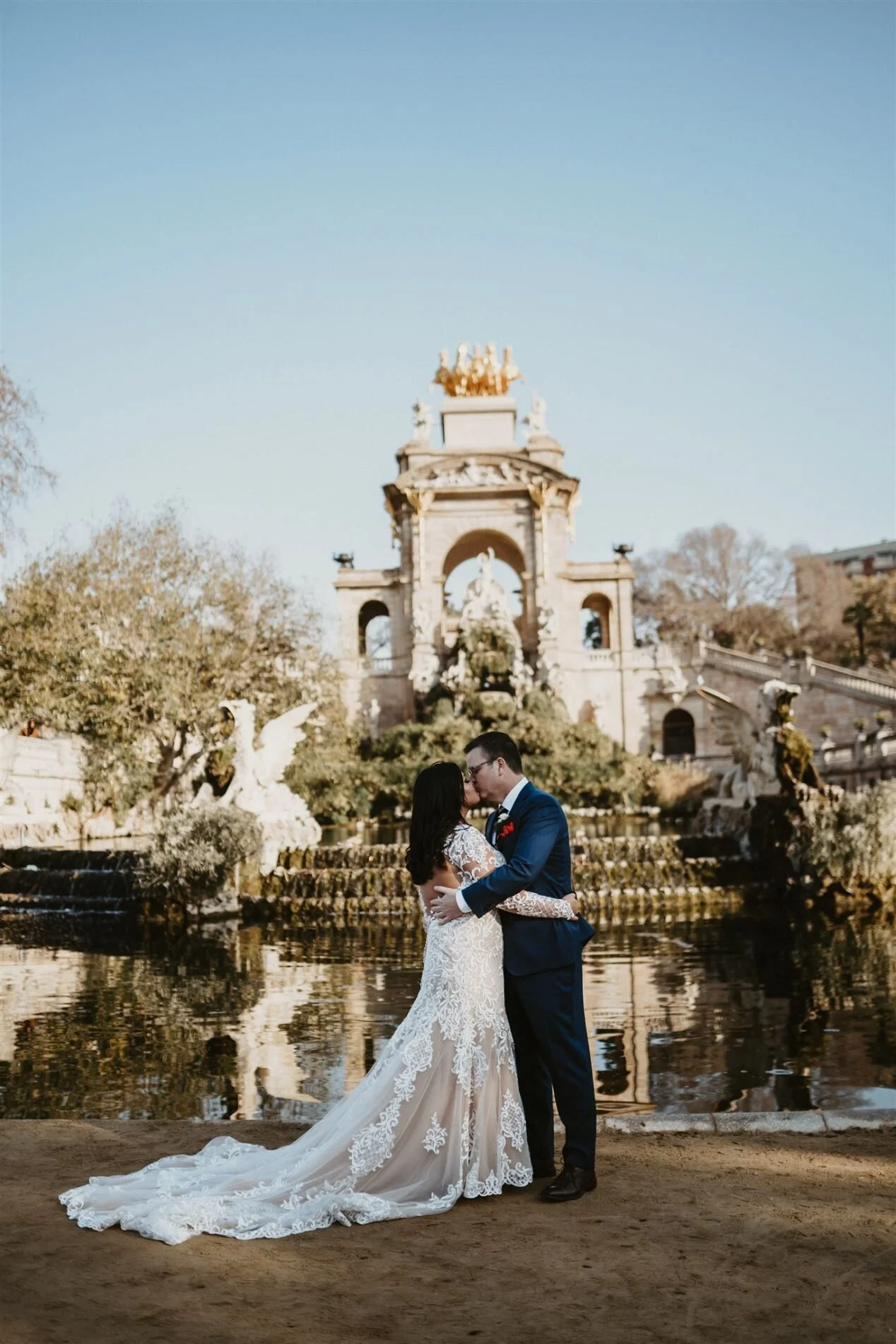 A bride and groom embrace for a wedding photo by a fountain with statues and a grand monument in the background. The bride is in a white lace wedding gown with a train, and the groom is in a dark suit. The scene is outdoors and appears to be in a par