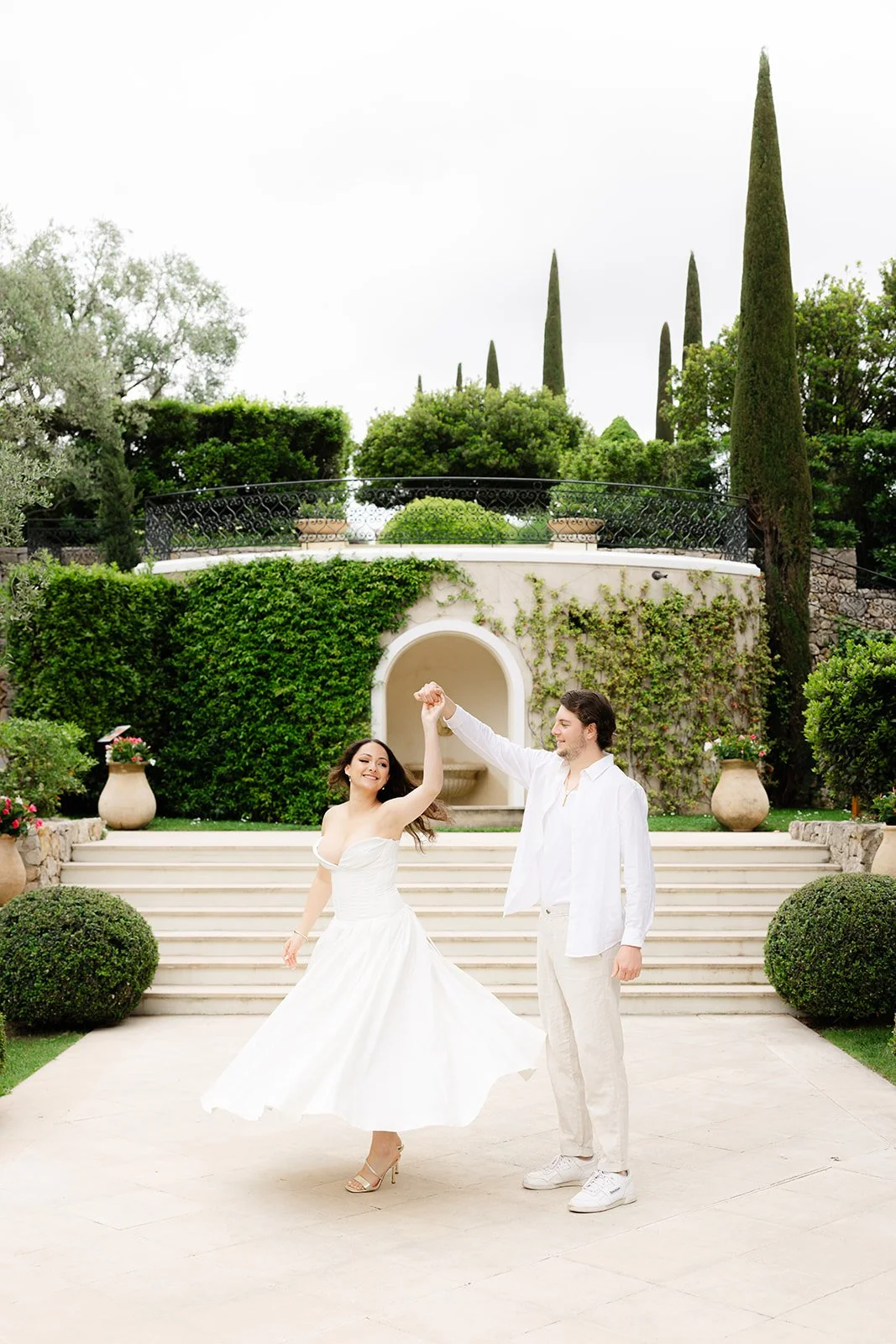 A couple dancing in wedding attire outdoors with greenery, stairs, and architectural elements in the background.