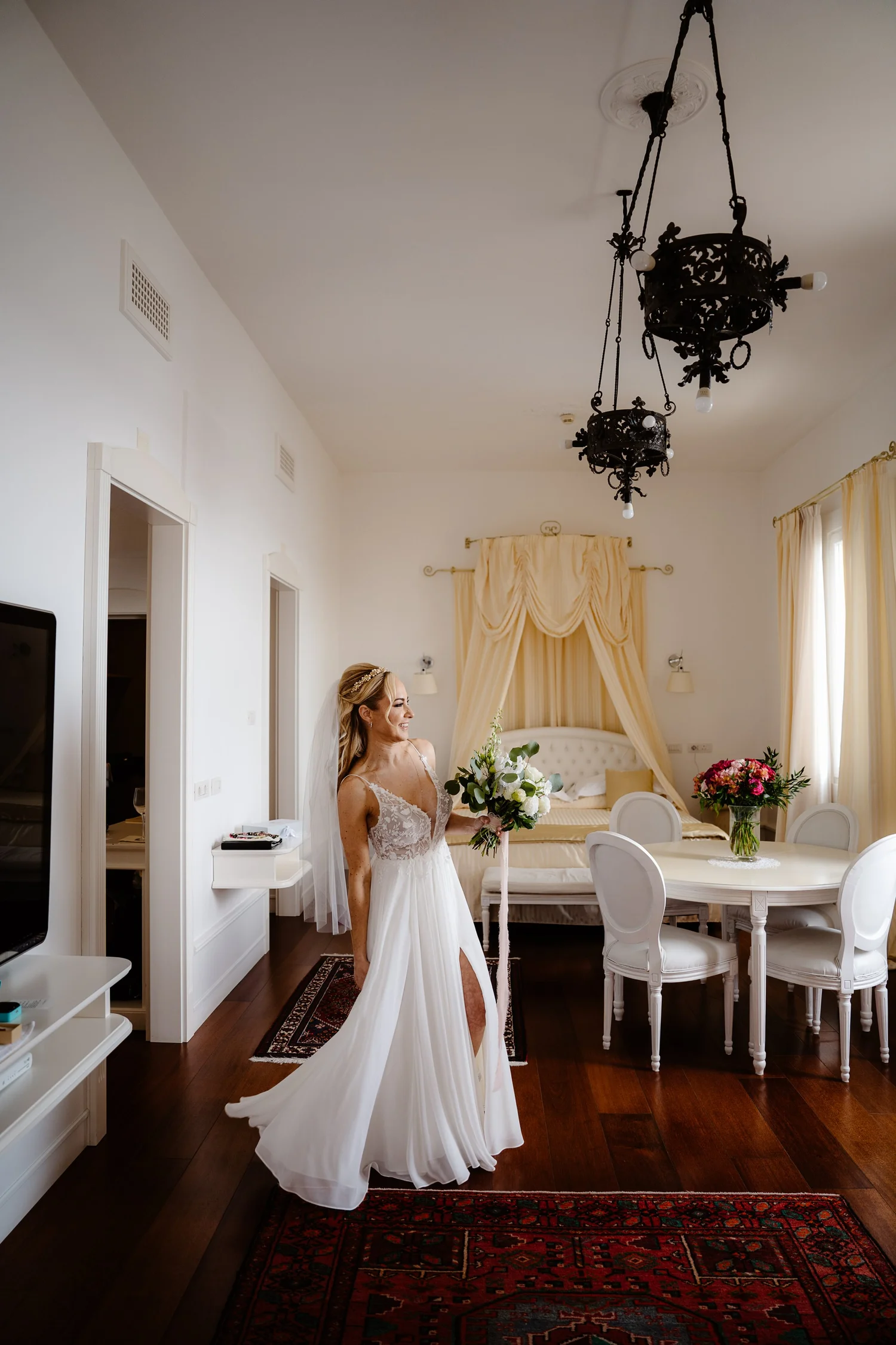 A bride in a white wedding dress holding a bouquet of white flowers, standing in a well-lit elegant room with high ceilings, ornate black chandeliers, and classic decor, including cream-colored curtains and a dark wooden floor.