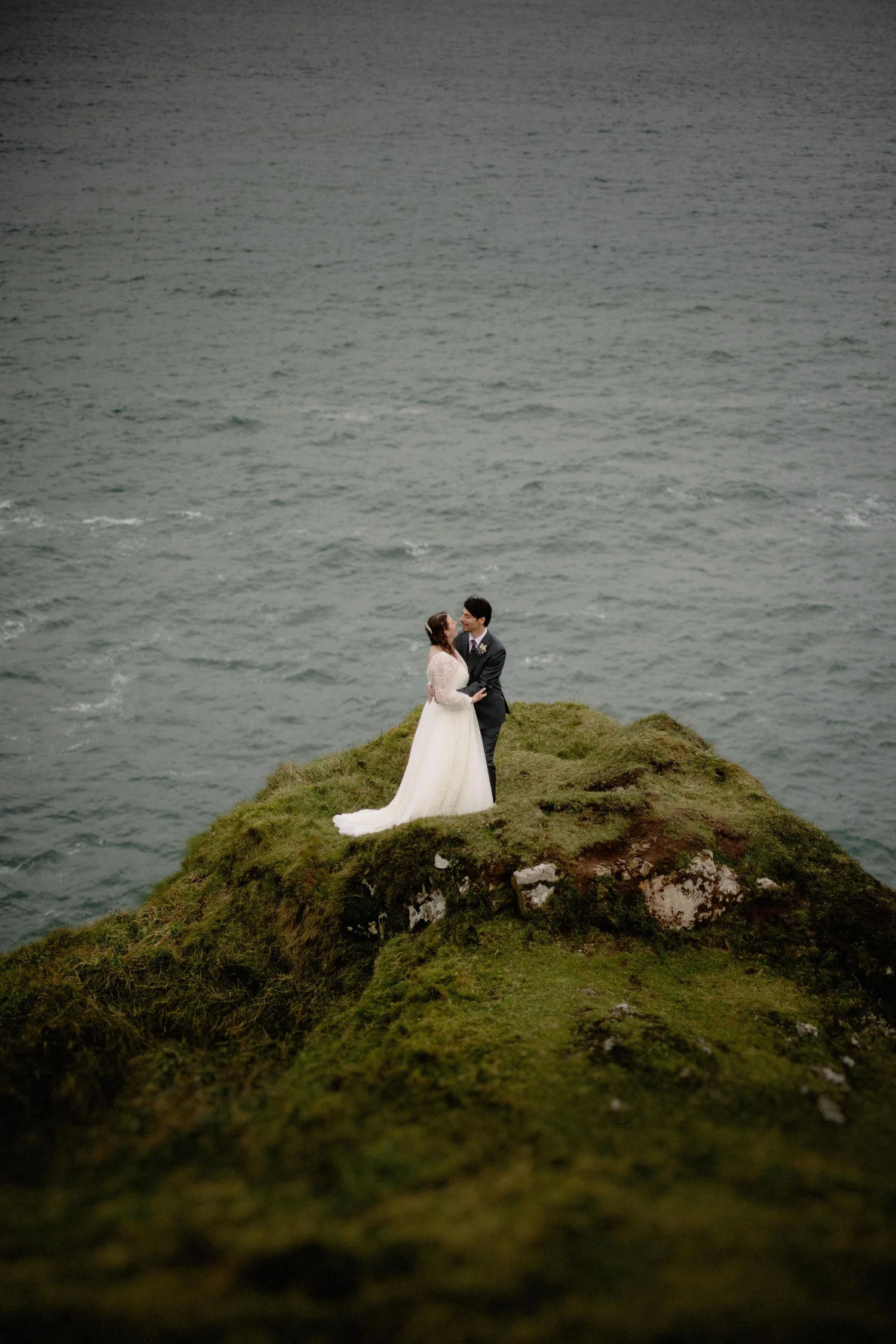 A bride and groom standing on a grassy cliff edge near the ocean, holding hands and looking at each other. The bride wears a white gown and the groom a dark suit.