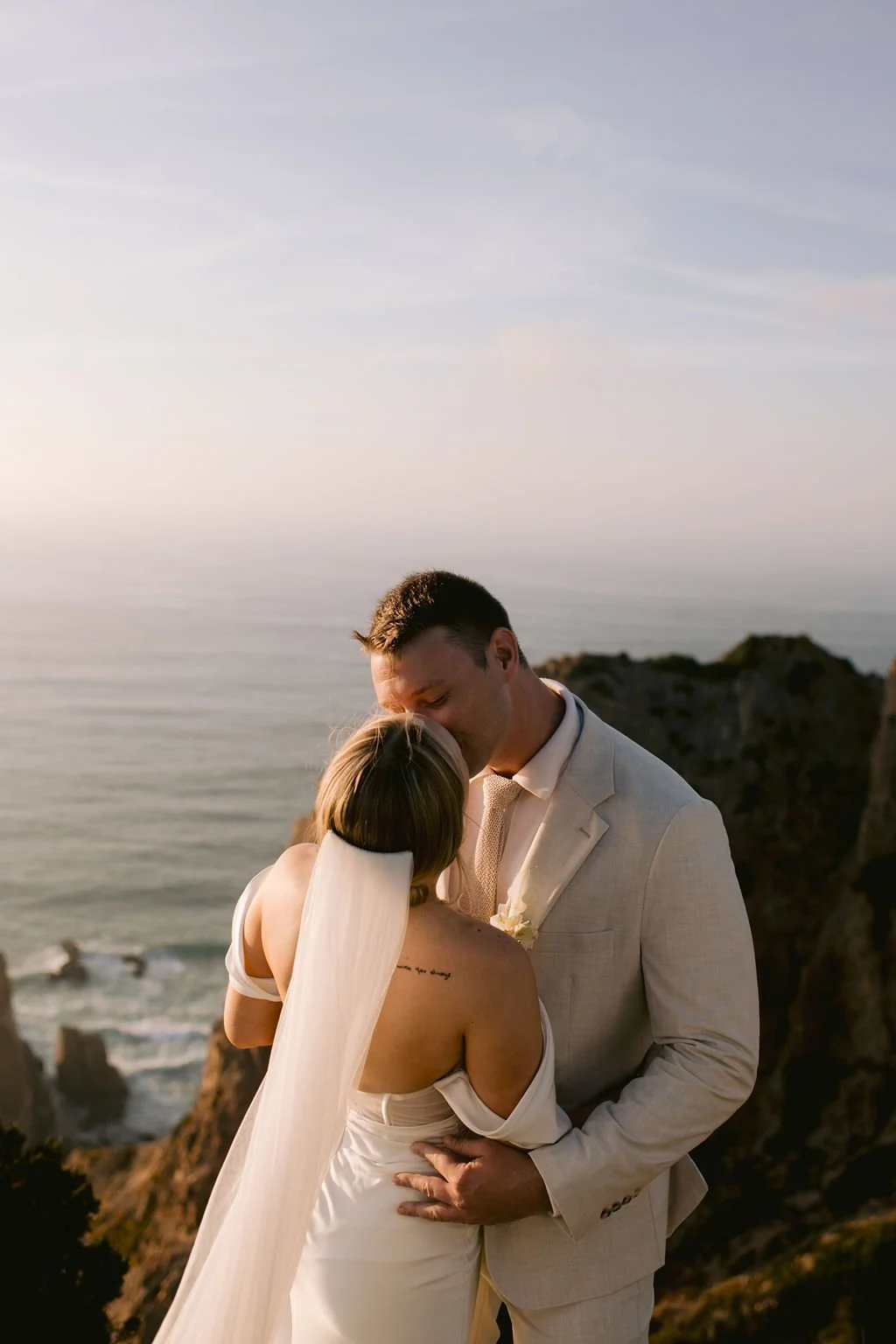 A newlywed couple sharing a kiss on a beach at sunset, with rocky cliffs and the ocean in the background.