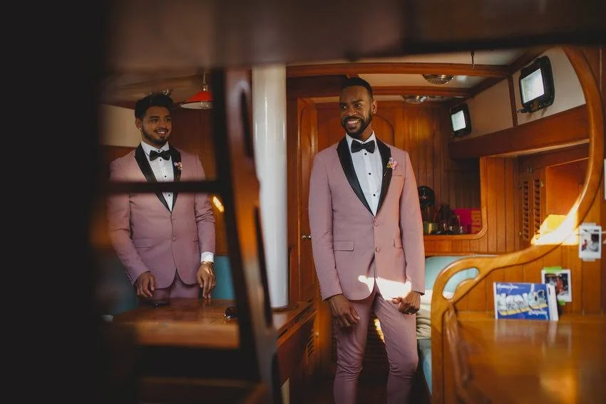 Two men in pink tuxedos with black lapels and bow ties, smiling inside a wooden room, with one looking into a mirror and the other standing near a counter.