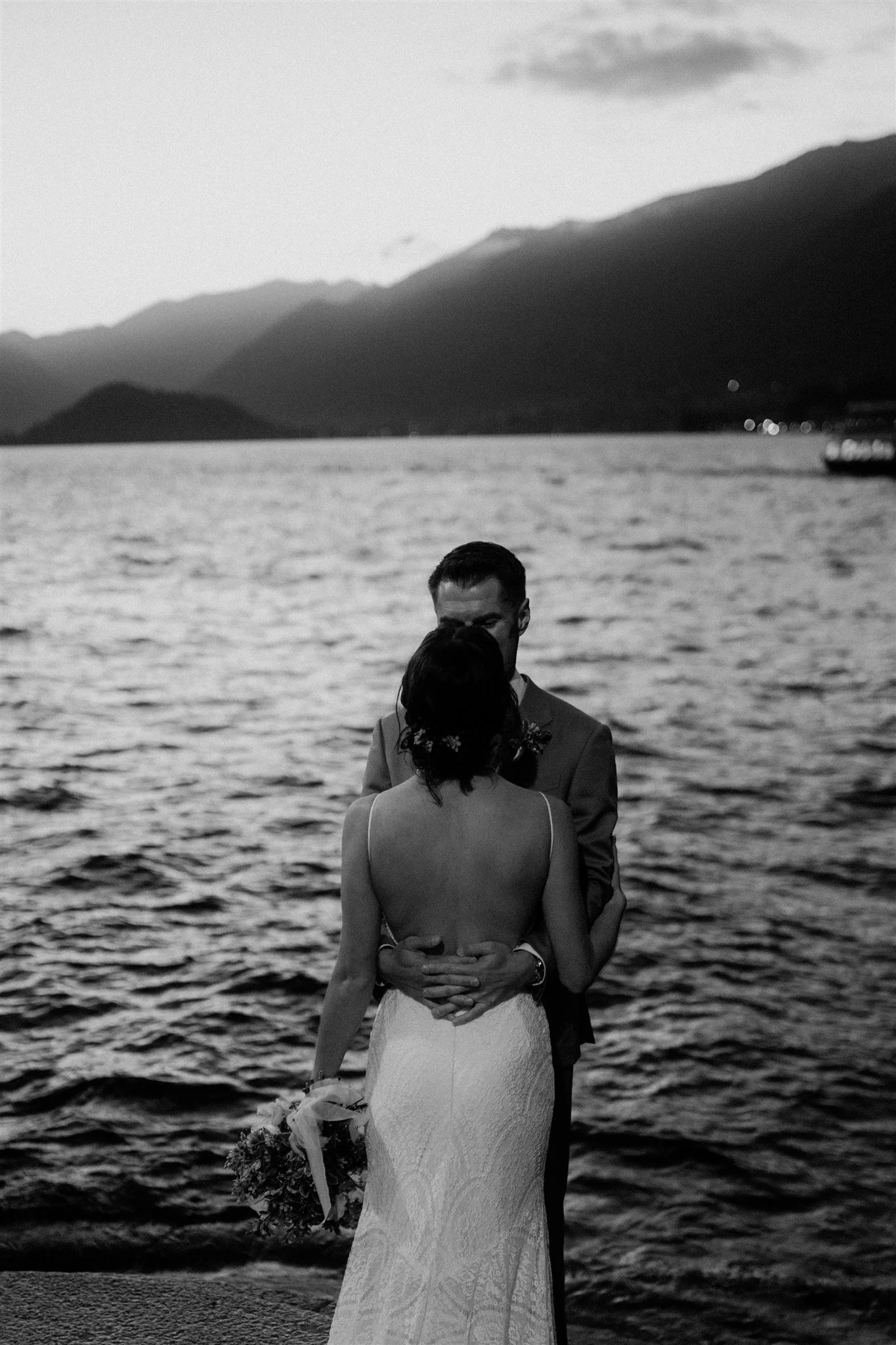 A black and white photo of a couple embracing near a body of water with mountains in the background. The woman is wearing a sleeveless dress and holding a bouquet of flowers. The man is wearing a suit, and they are kissing.