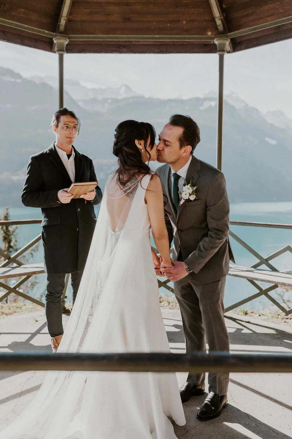 A bride and groom kiss during their wedding ceremony on a gazebo overlooking a lake and mountains, with an officiant holding a book nearby.