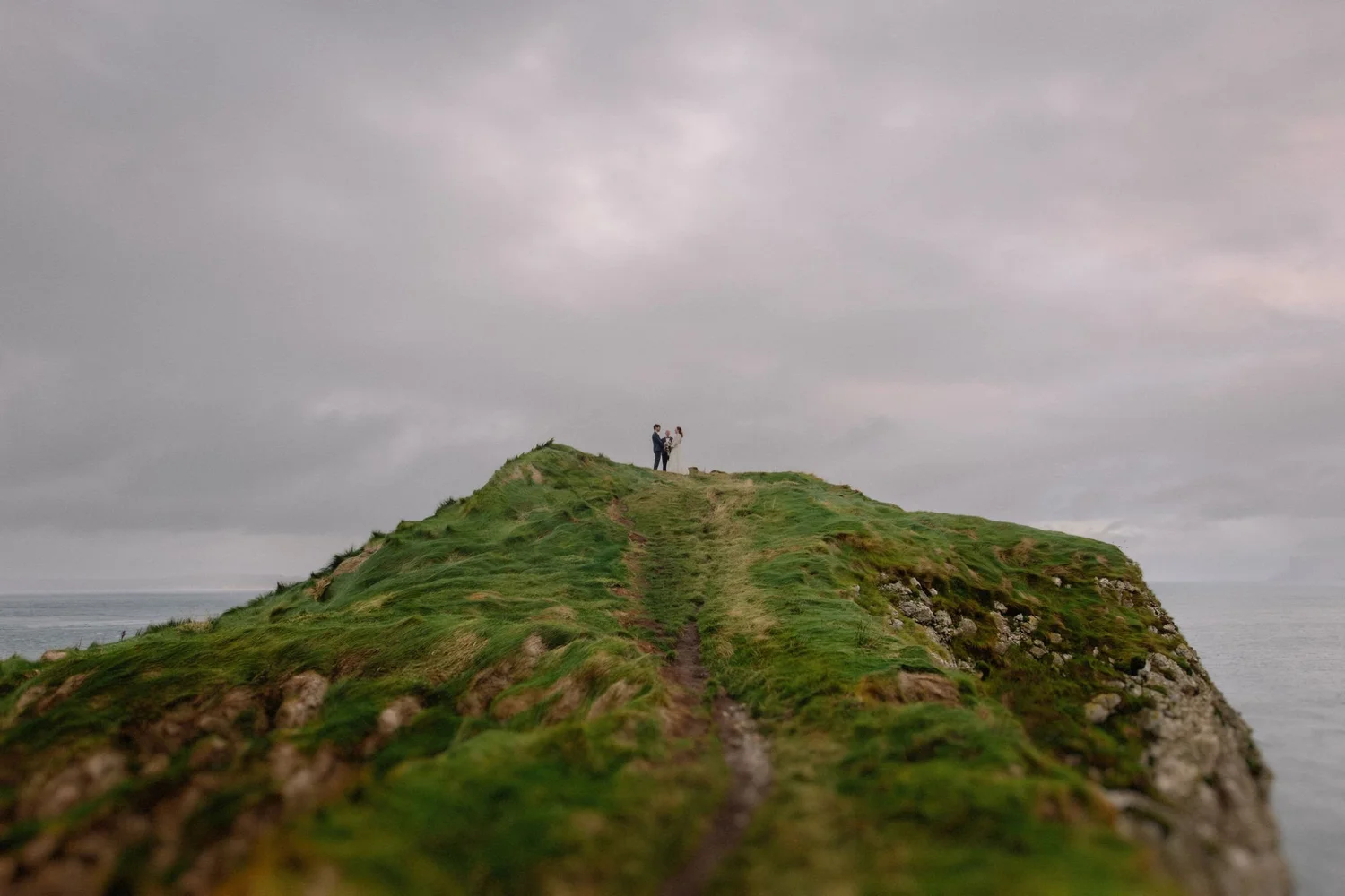 A bride and groom standing on a grassy hilltop overlooking the ocean, under a cloudy sky.