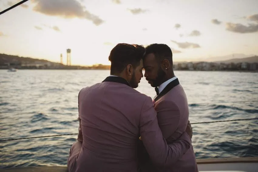 Two men in pink suits embracing on a boat at sunset with water and cityscape in the background.