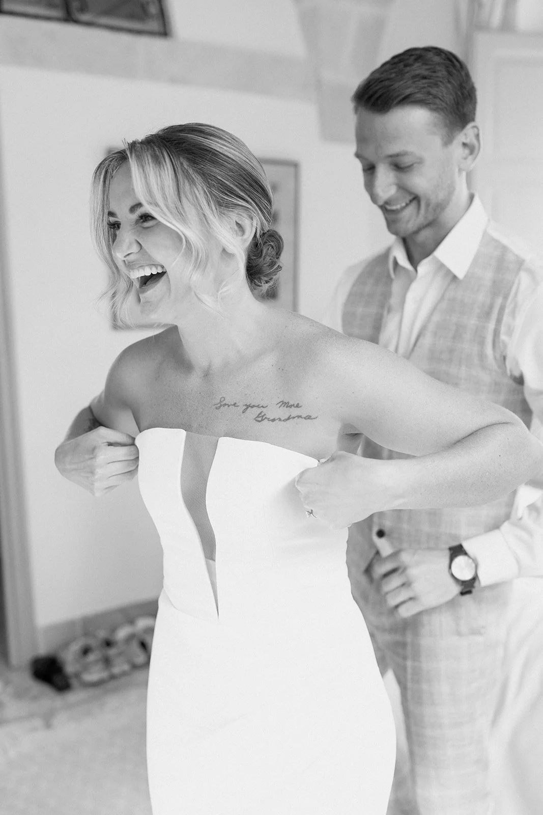 A woman in a wedding dress excitedly adjusts the front of her dress, revealing a tattoo on her chest that says "Love you Mom Grandma." She is smiling and laughing, with a man behind her helping her, both in a room with a light-colored wall.