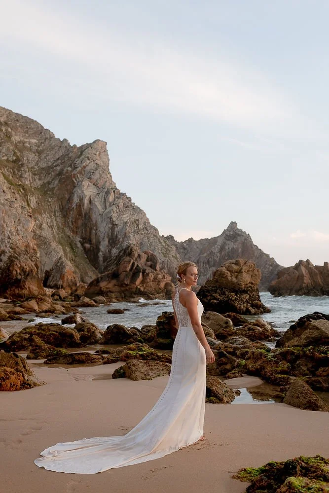 A woman in a white wedding dress standing on a rocky beach with cliffs and ocean in the background during sunset.