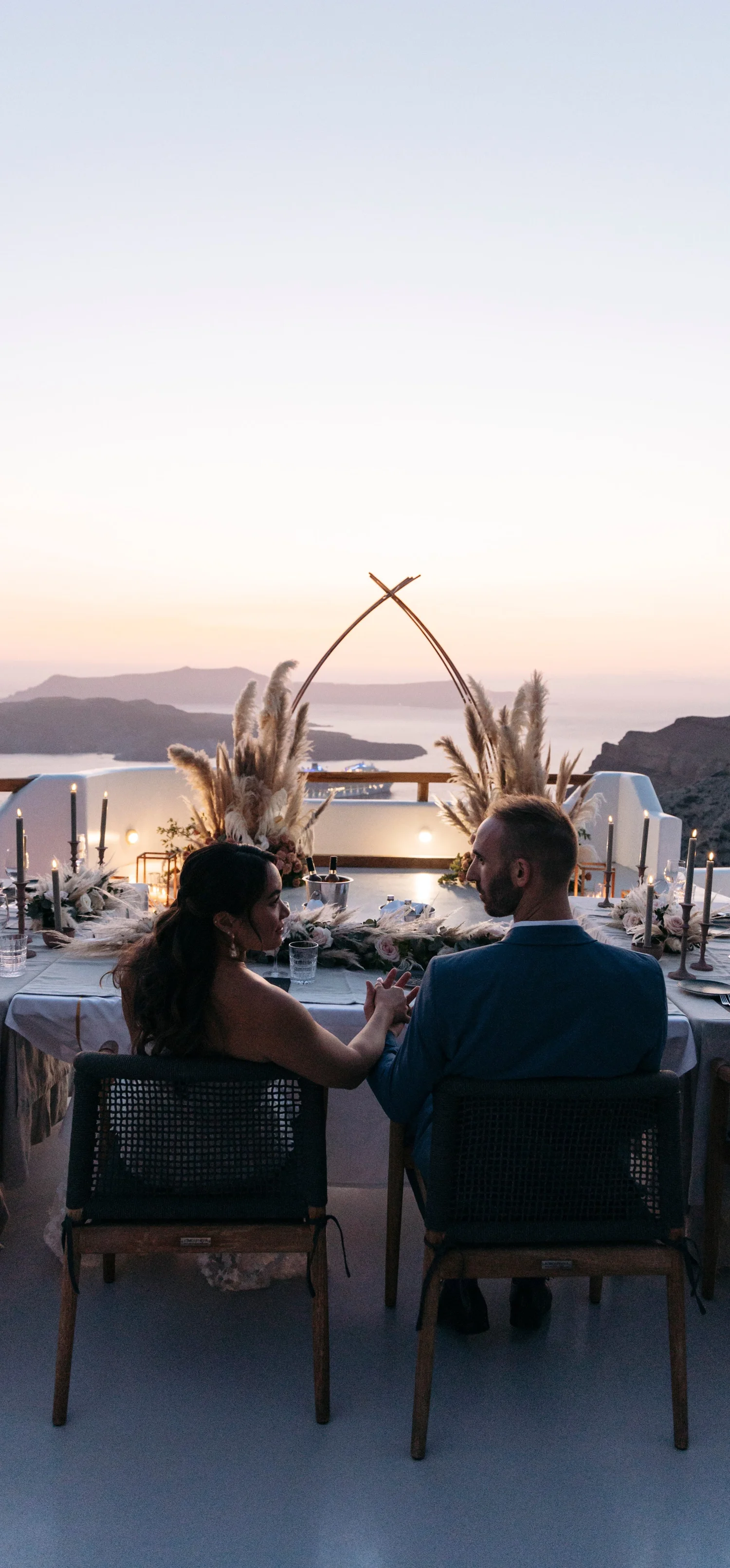 A romantic outdoor dinner table set on a terrace overlooking the ocean at sunset, decorated with candles, flowers, and pampas grass, with a couple holding hands and enjoying the view.