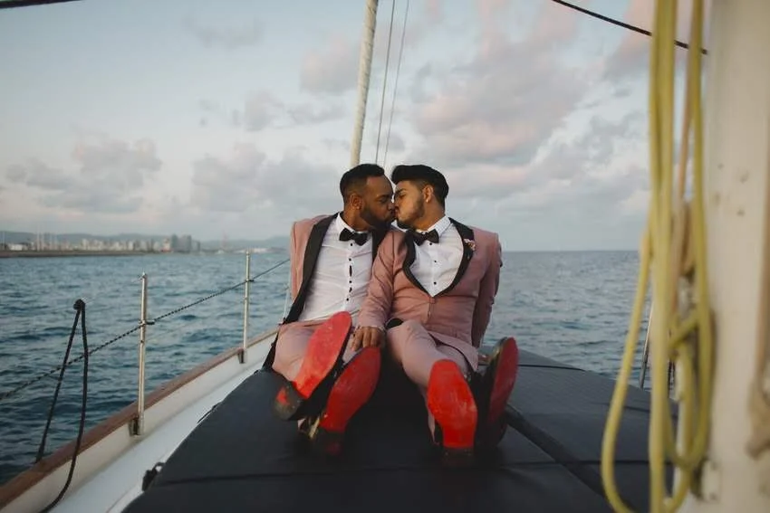 Two men dressed in pink suits and black bow ties sitting close together on a boat at sea, about to kiss, with city skyline in the distance and a cloudy sky overhead.