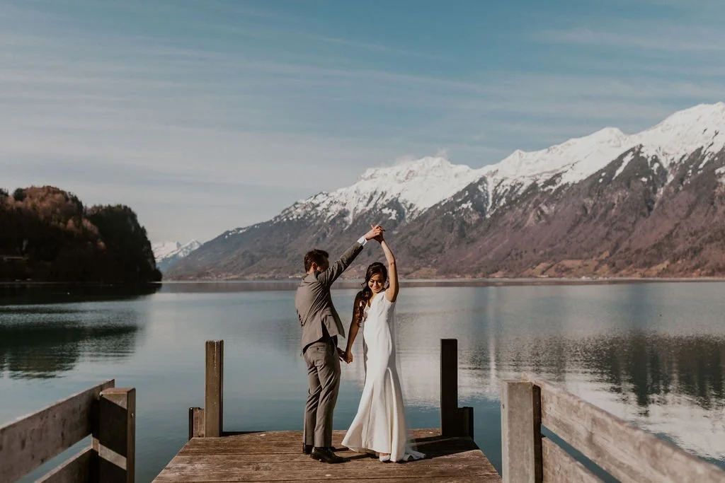 A couple in wedding attire dancing on a wooden dock by a calm lake, with snow-capped mountains in the background.